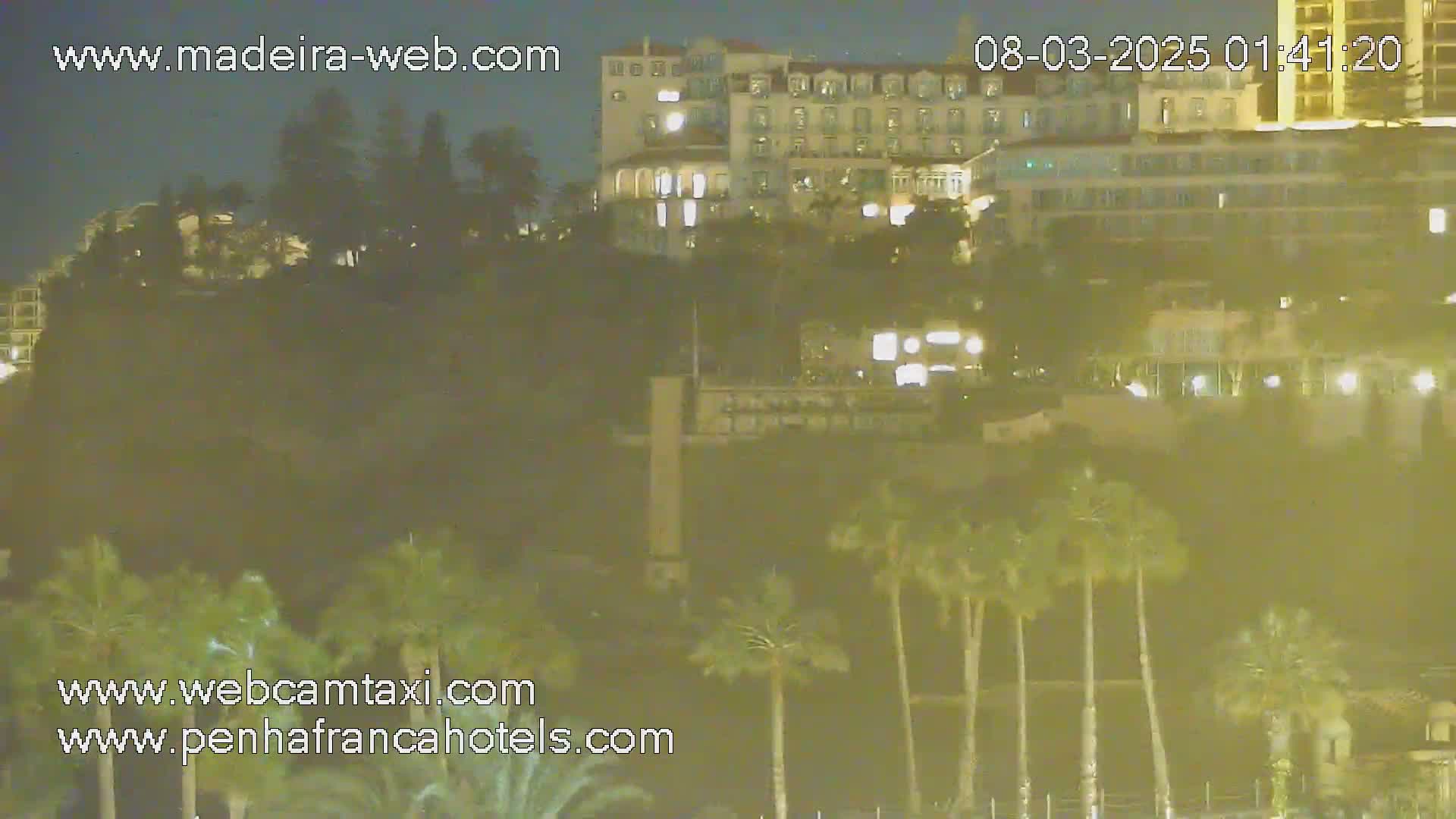 A nighttime long shot shows a large building illuminated against a dark hill dotted with palm trees under a clear sky.