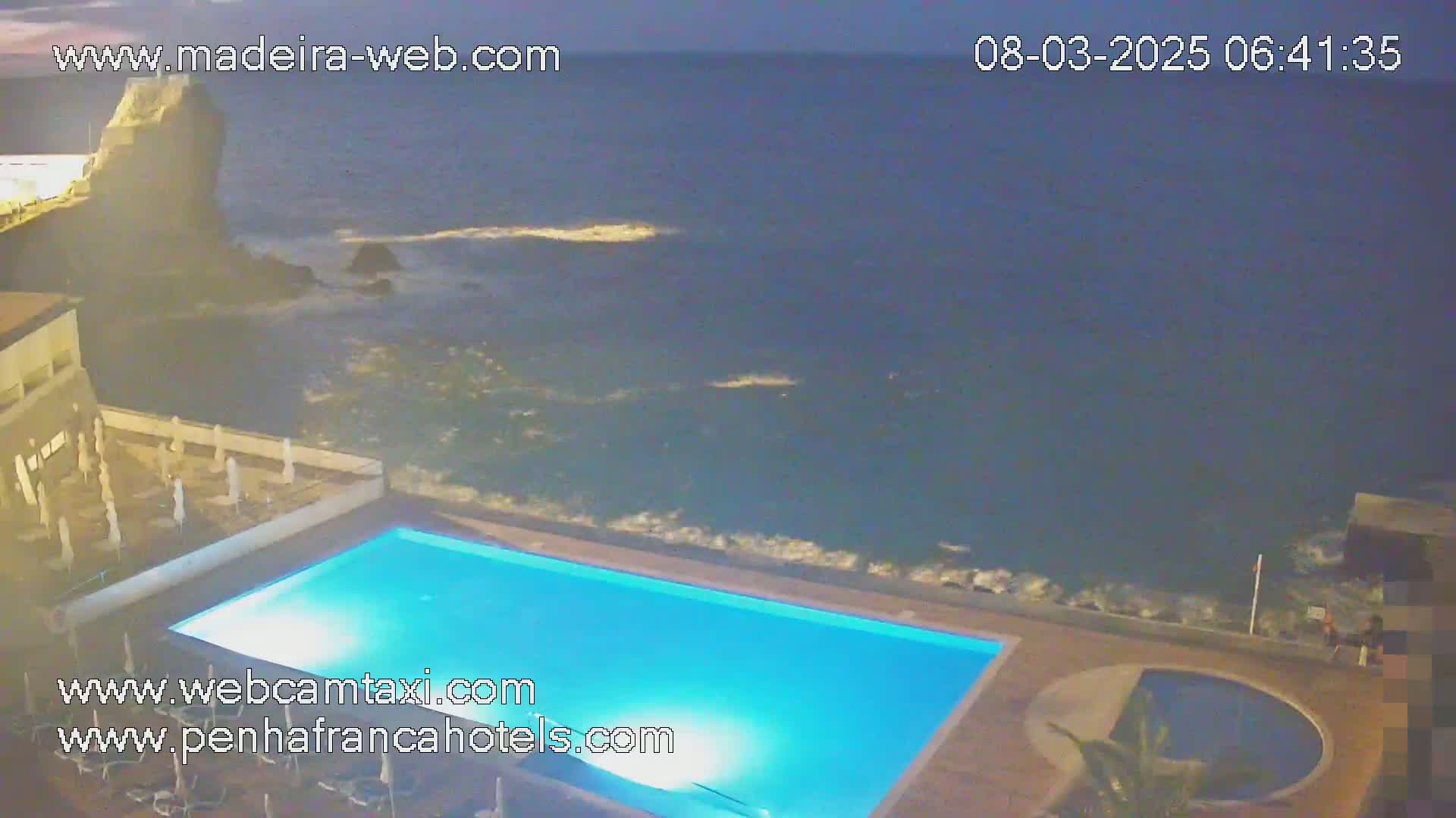 A nighttime view of a rectangular swimming pool next to a dark ocean with small waves crashing against the rocks, under a clear, dark sky.