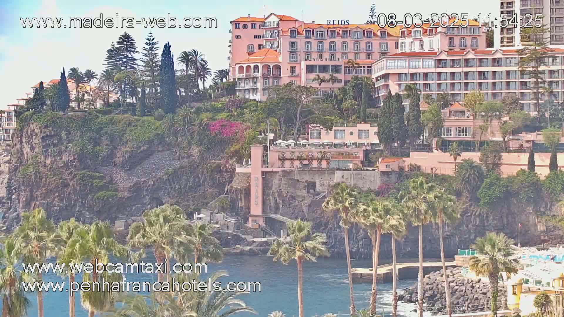 A pink hotel complex sits atop a rocky cliff overlooking a calm ocean dotted with palm trees under a sunny sky.