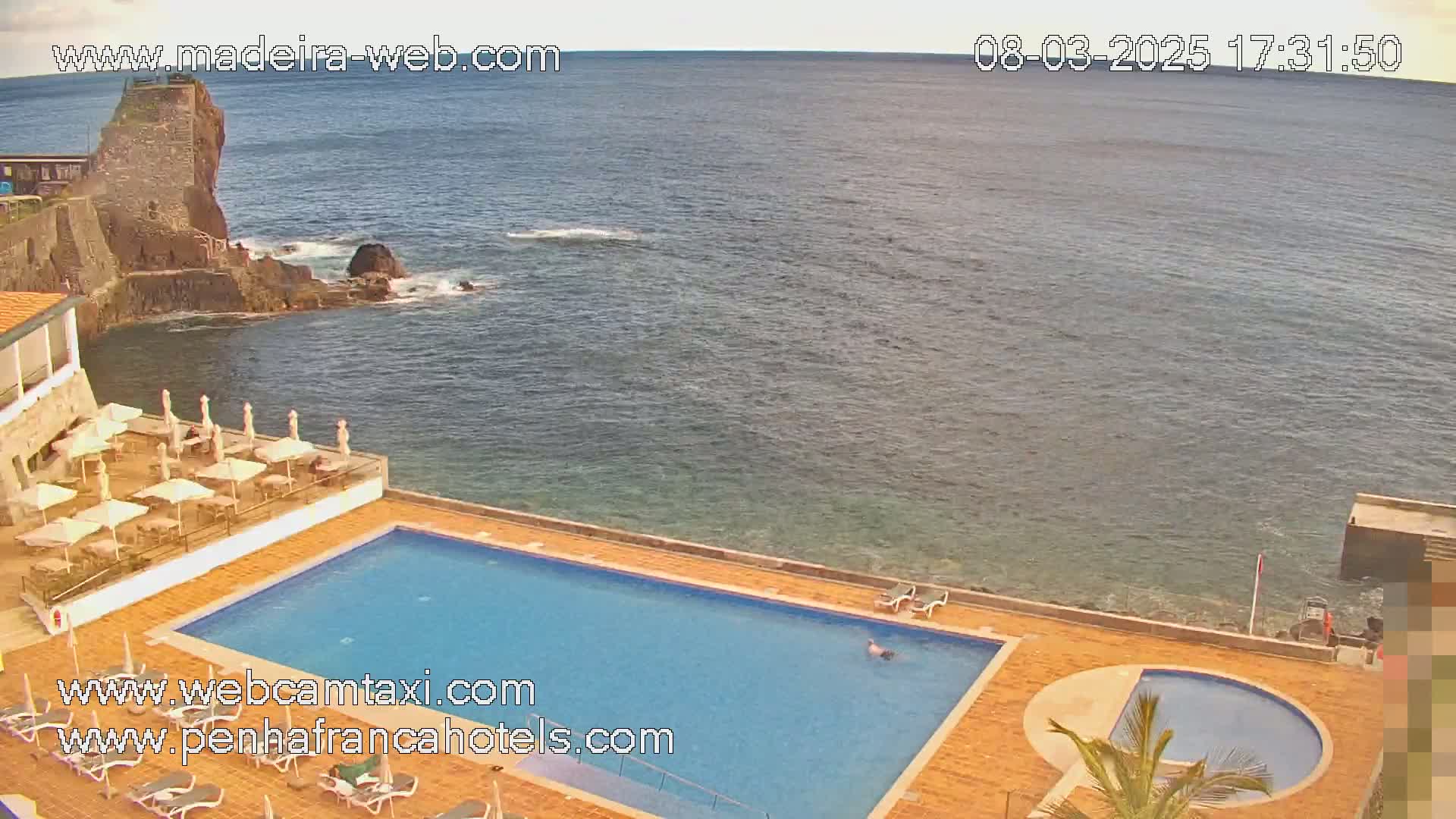 An outdoor swimming pool and smaller circular pool are situated next to a patio with umbrellas and chairs, overlooking a calm ocean and rocky coastline on a mostly clear day.