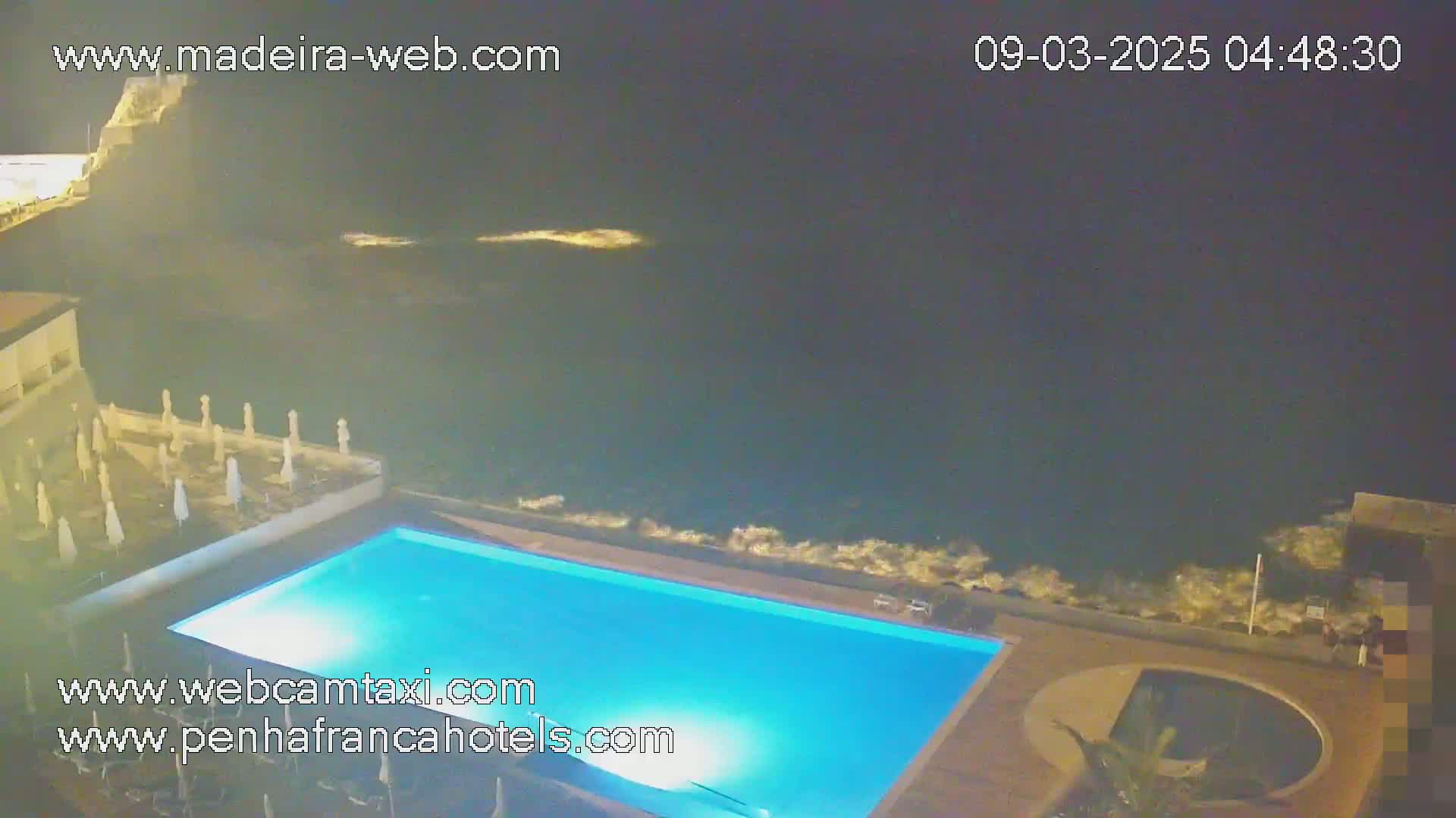 A nighttime view of a lit, rectangular swimming pool situated next to a dark ocean with some small waves.