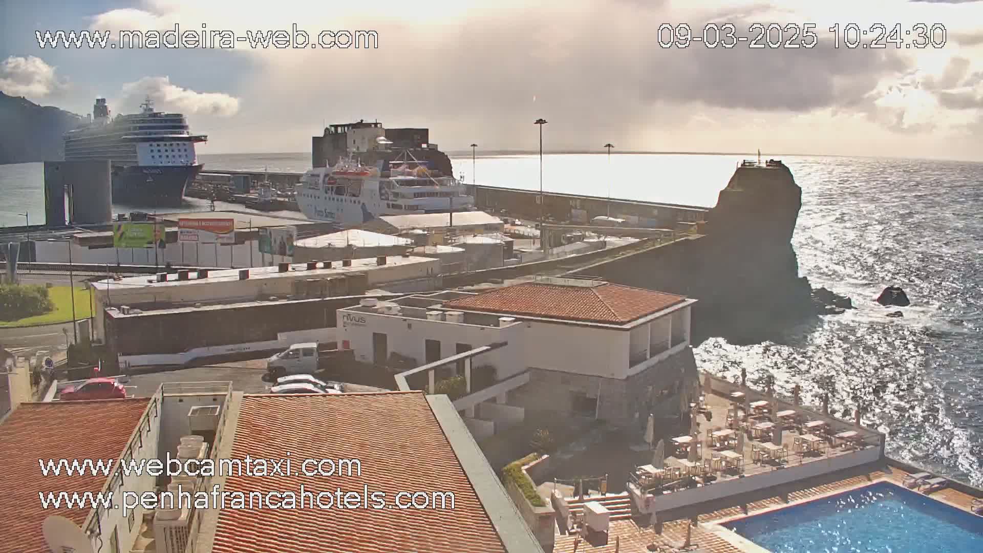 A harbor with two docked ships is visible from an elevated viewpoint overlooking a hotel with an outdoor pool and patio under partly cloudy skies.