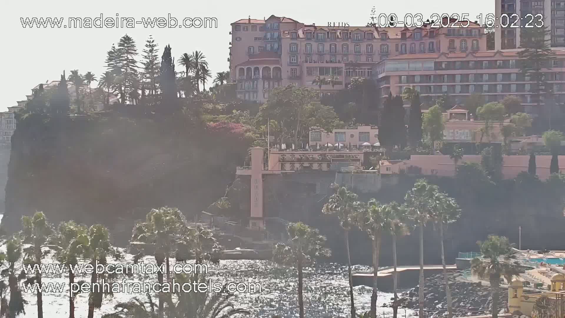 A hazy view shows a rocky coastline with hotels and lush vegetation, overlooking a wavy ocean.