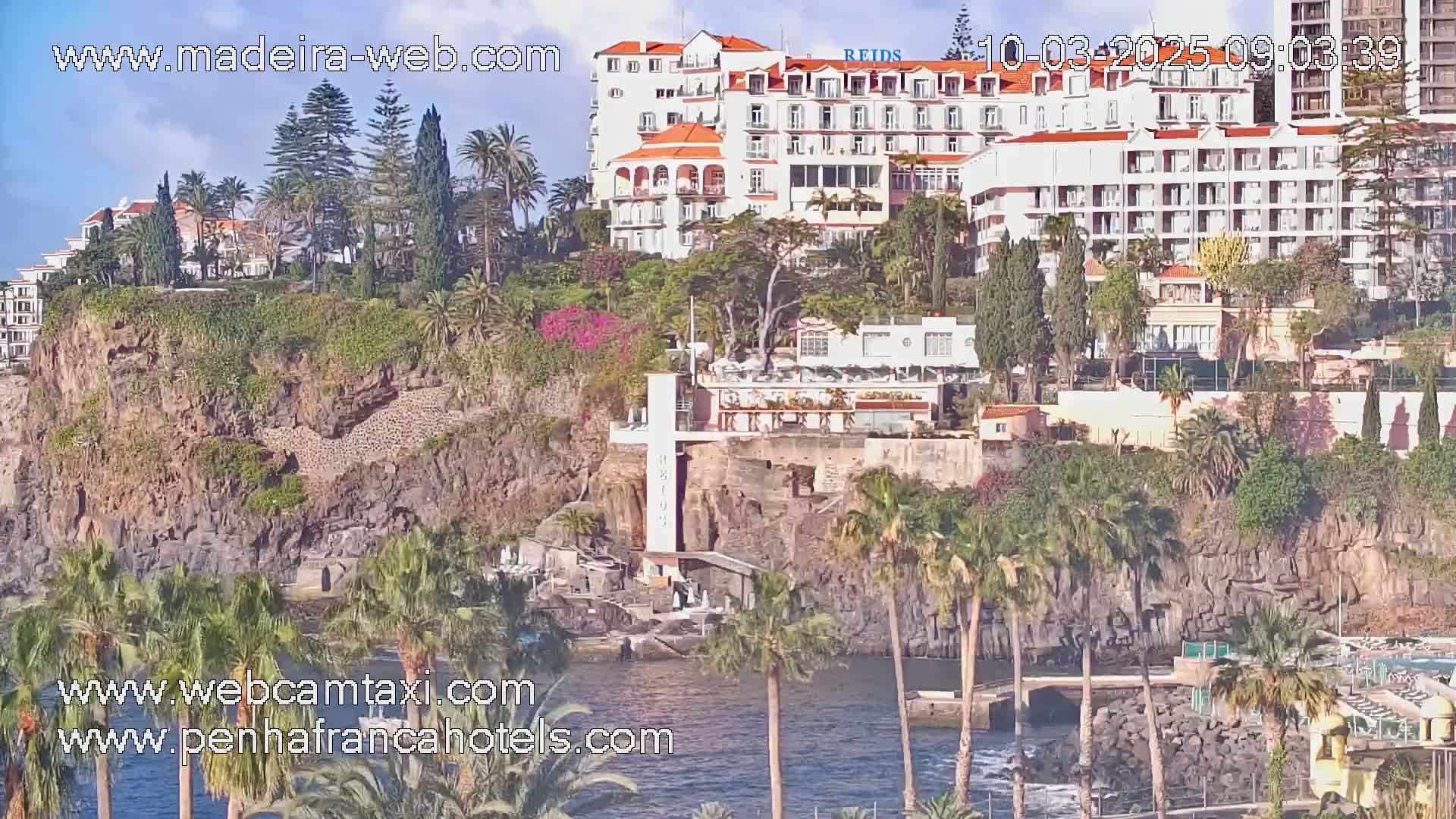 A large white hotel sits atop a rocky cliff overlooking a calm body of water, surrounded by lush tropical vegetation under a mostly sunny sky.