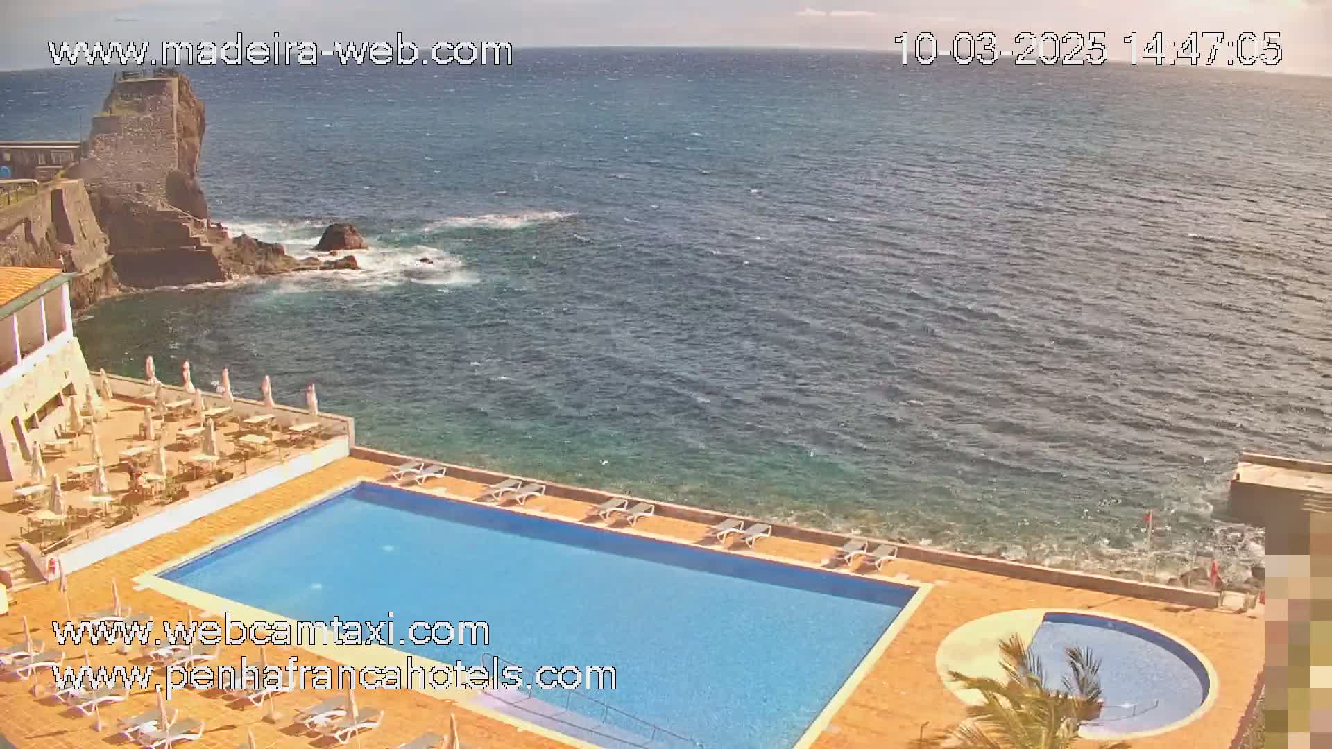 A hotel pool and patio area overlook a rocky coastline and ocean under a sunny sky.