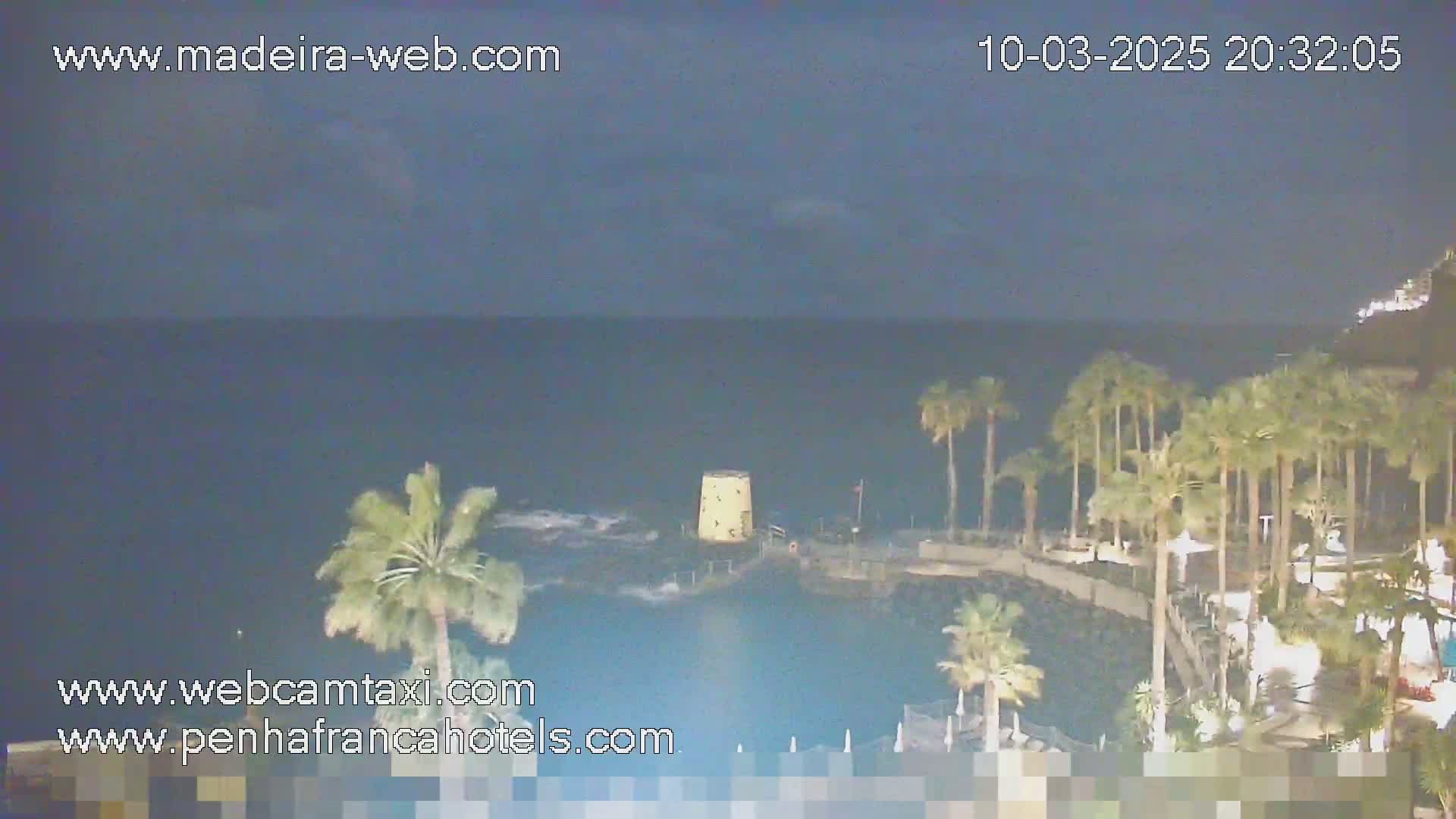 A nighttime coastal scene shows dark water, palm trees swaying in the breeze, and a small stone tower near the shore under a mostly clear, dark sky.