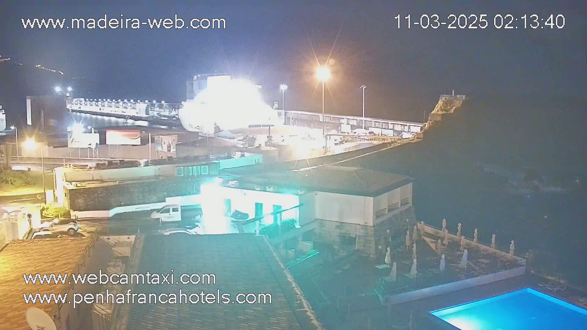 A nighttime, aerial view shows a large ship docked at a port, a building with a pool, and surrounding structures under partly cloudy skies.