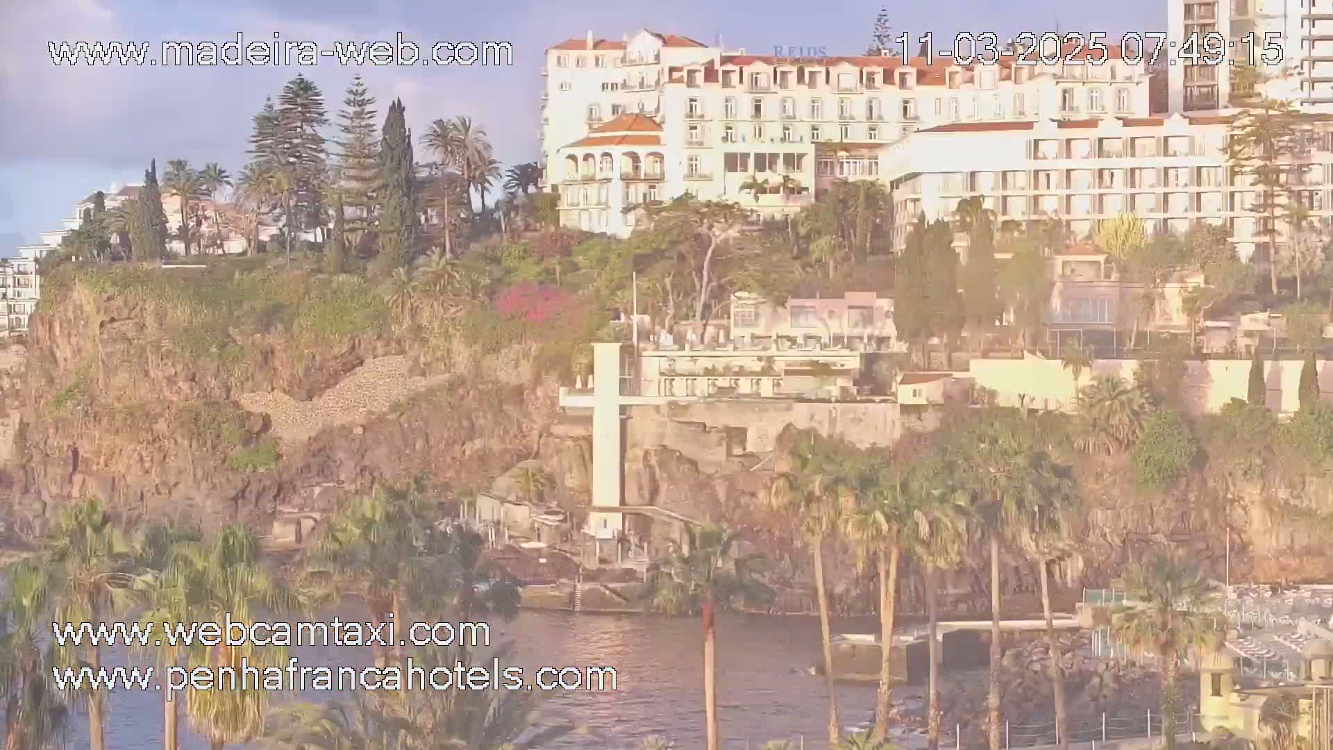 A coastal scene with a large white hotel,  palm trees, and rocky cliffs overlooks a calm body of water under a mostly clear sky.