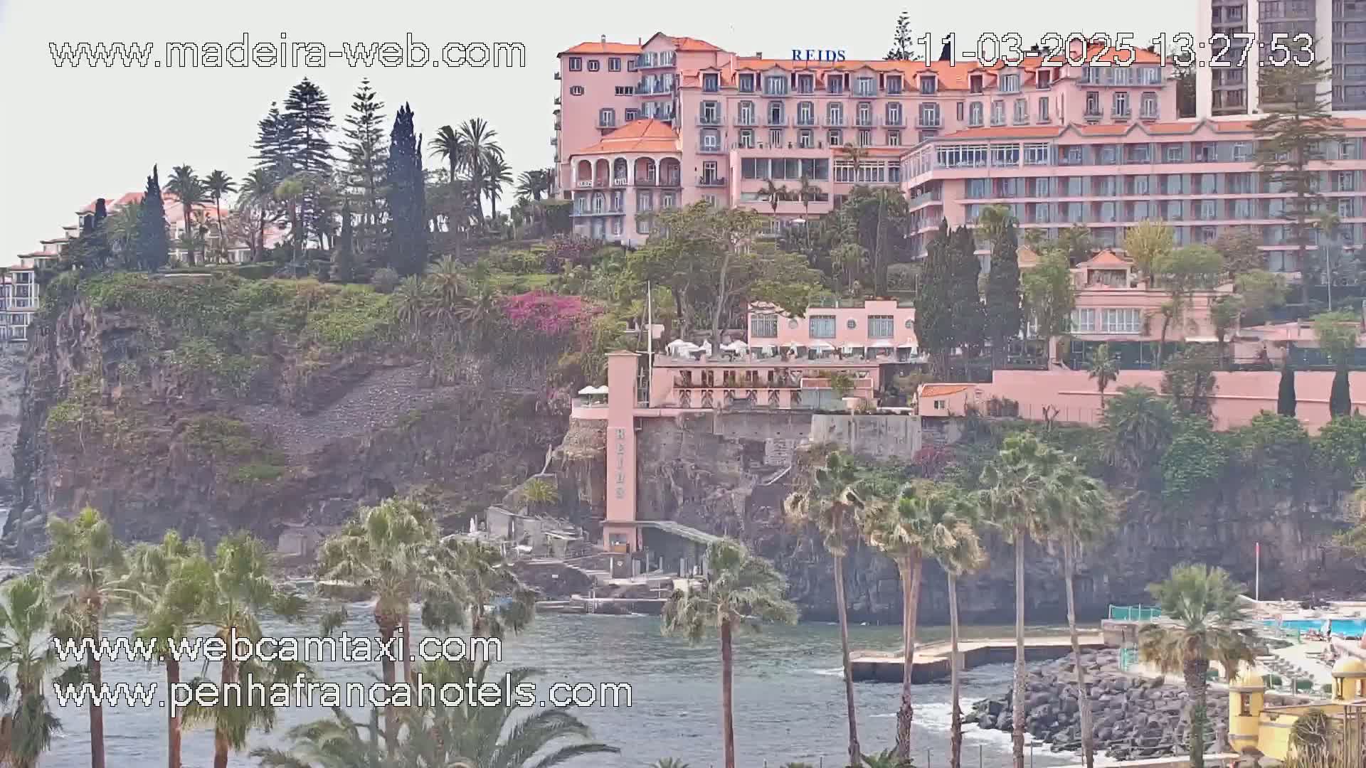 A pink hotel complex sits on a cliff overlooking a calm body of water, surrounded by lush greenery and palm trees under an overcast sky.