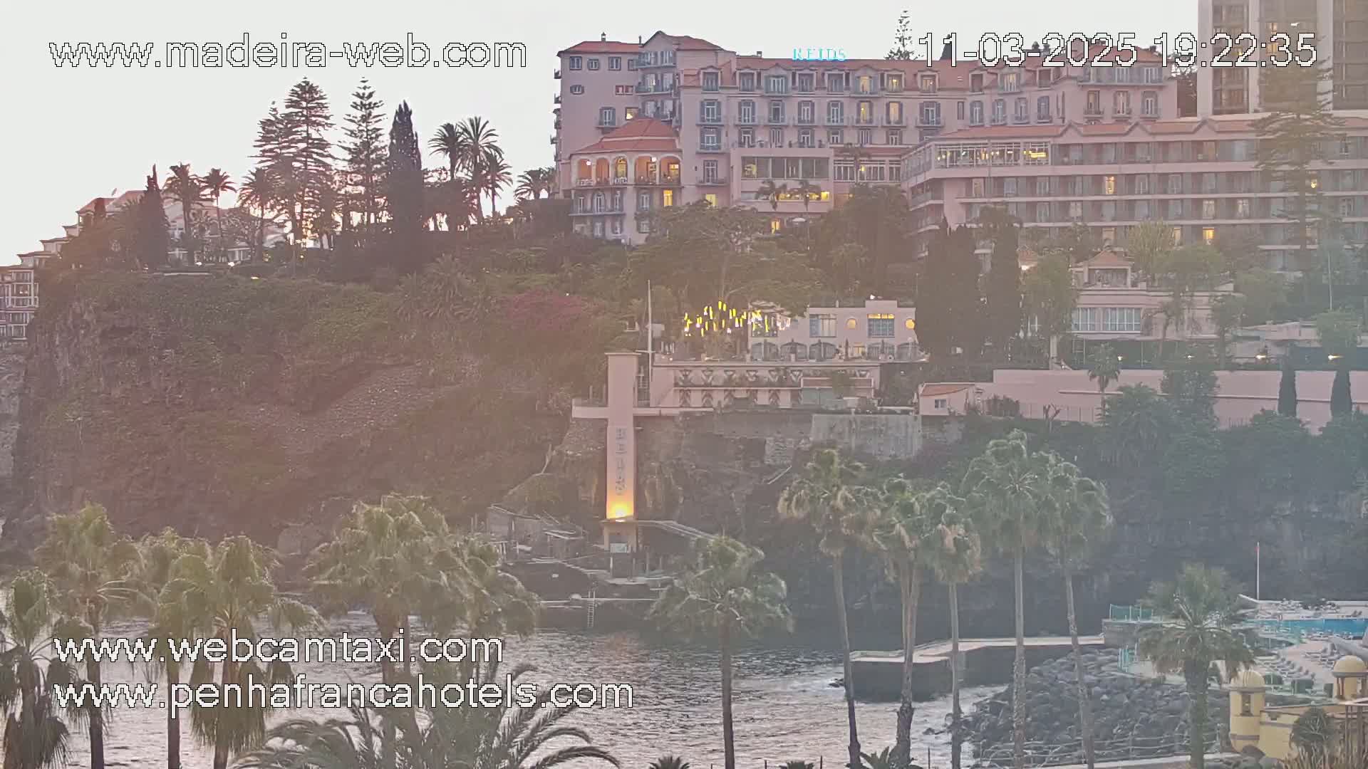A coastal cliffside scene at sunset shows several large hotels and buildings with palm trees, lush vegetation, and a rocky shoreline with a calm ocean.