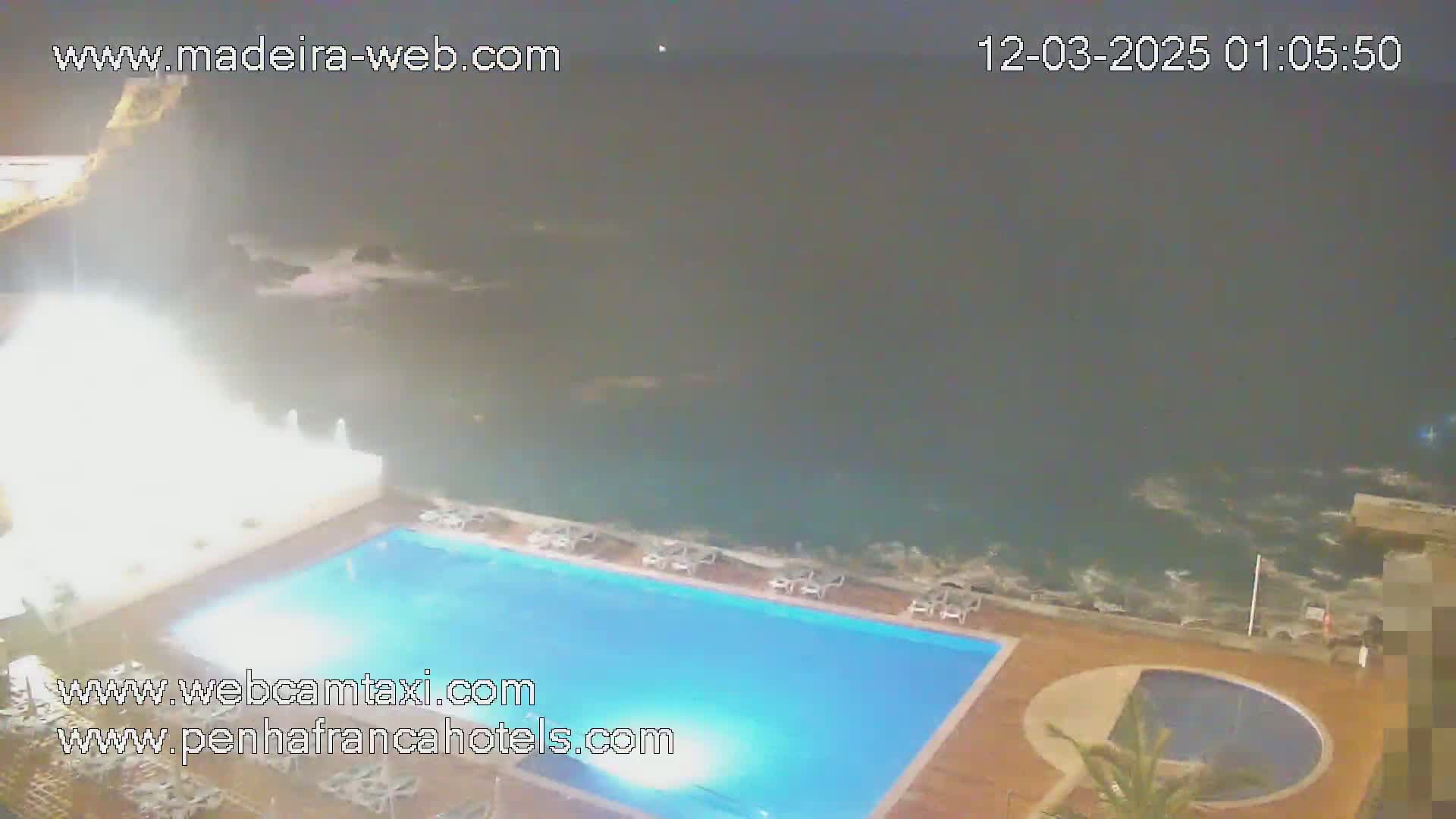 A nighttime aerial view shows a rectangular swimming pool next to a smaller, round pool, situated on a deck overlooking a dark, wavy ocean.