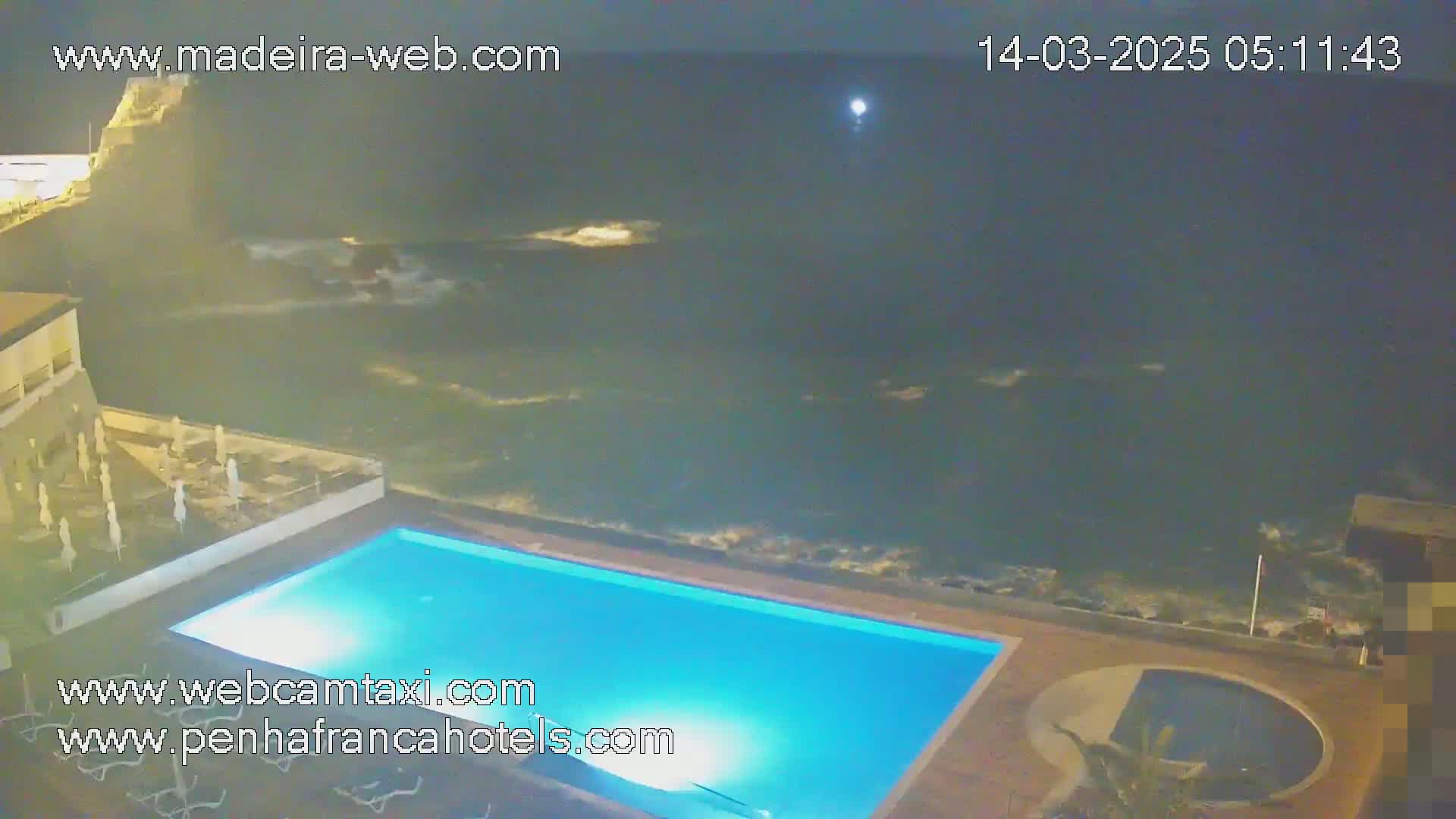 An illuminated swimming pool sits adjacent to a rocky coastline under a dark, nighttime sky with a bright moon.