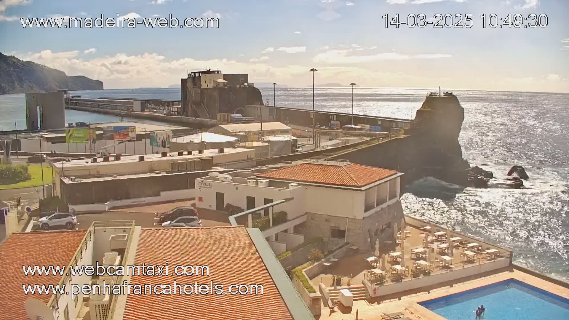 An oceanside hotel with an outdoor pool overlooks a harbor and rocky coastline under a mostly sunny sky.