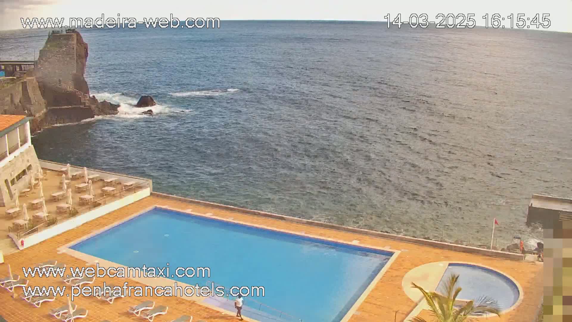 An outdoor swimming pool and a smaller adjacent round pool are situated on a patio overlooking a calm ocean next to dark volcanic rock formations.