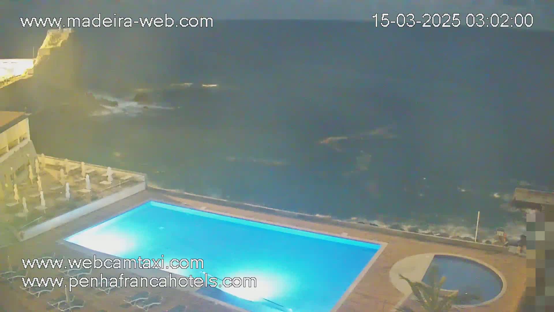 A nighttime view of a rectangular swimming pool next to a rocky ocean shore under a dark, cloudy sky.