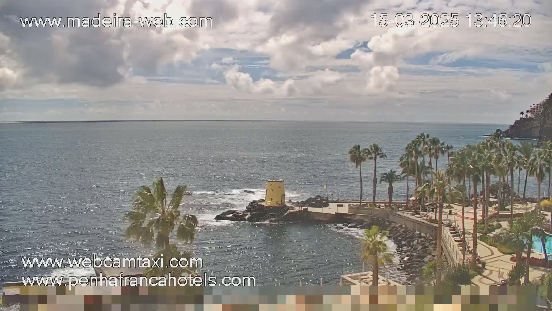 A partly cloudy day shows a rocky coastline with palm trees, a small yellow structure on a rock outcropping in the ocean, and calm ocean waters.
