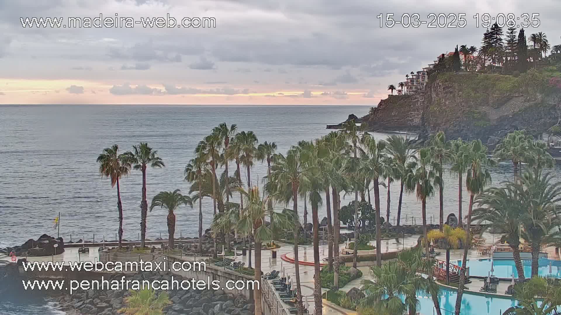 A resort with palm trees and swimming pools overlooks a calm ocean under a cloudy sky.