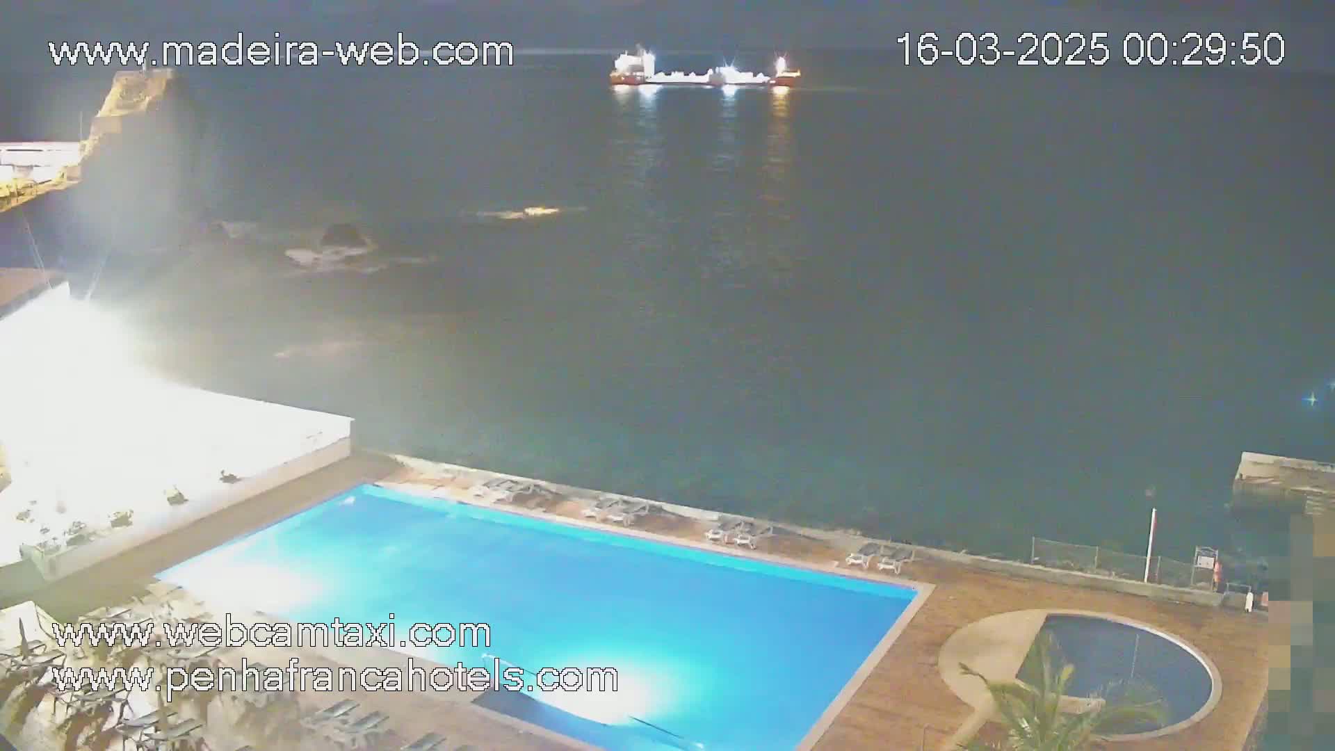 A nighttime view shows a rectangular swimming pool next to a dark ocean where a large ship is visible in the distance.