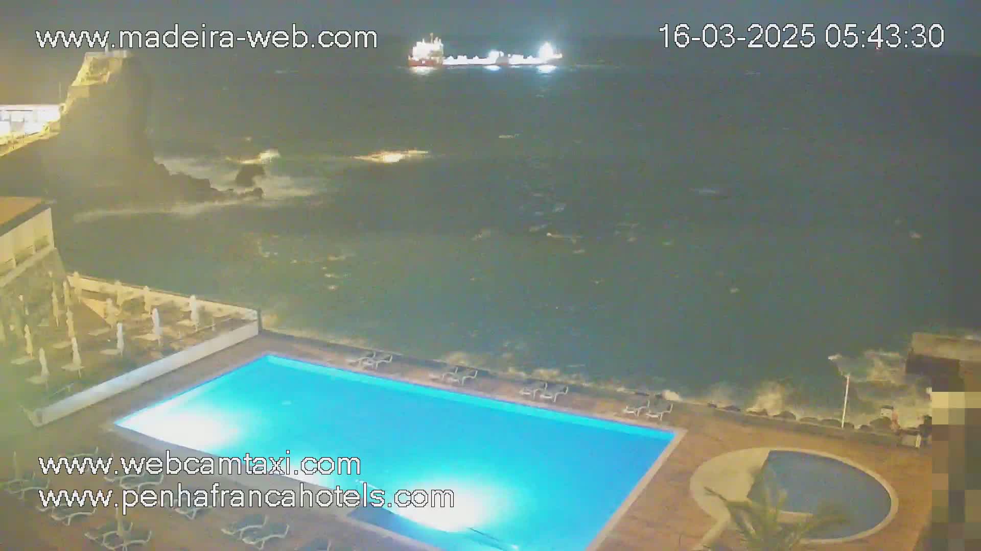 A nighttime view shows a swimming pool at a resort overlooking a dark, wavy ocean with a ship visible in the distance.