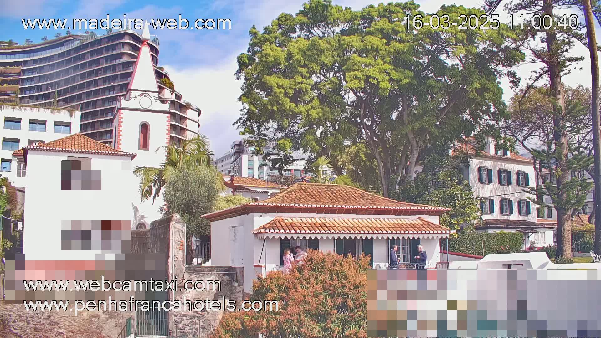 A partly sunny day shows several buildings, including a white building with a red tile roof and a taller white church-like structure, surrounded by lush green trees.