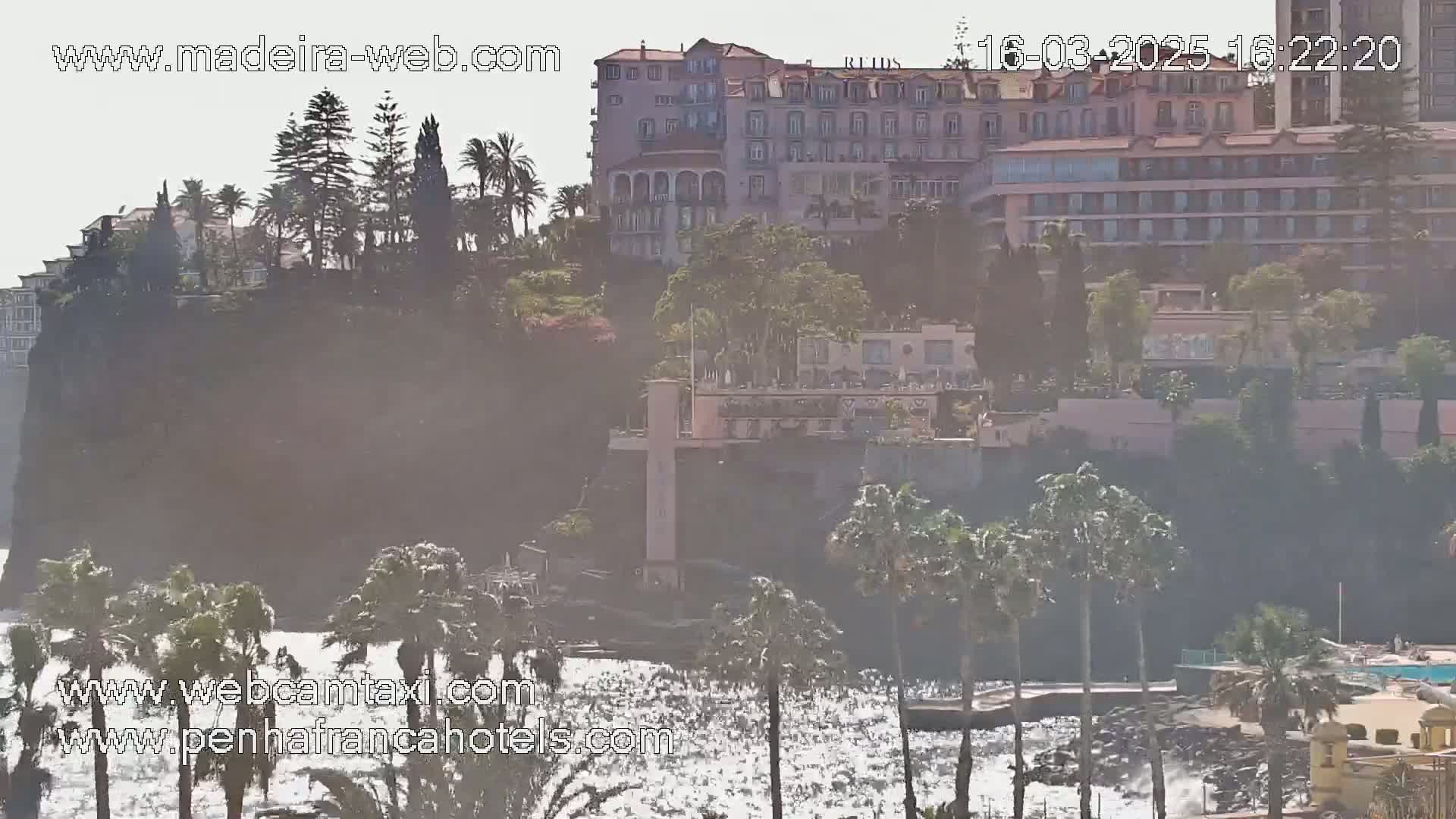 A hazy coastal scene shows several buildings on a cliff overlooking a rocky shore with a somewhat choppy sea.