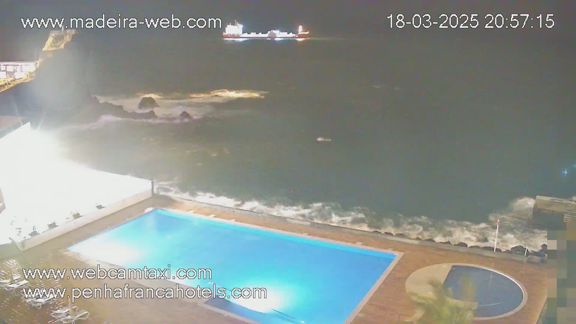 A nighttime view of a rectangular swimming pool next to a rocky ocean shore with moderate waves under a dark sky.