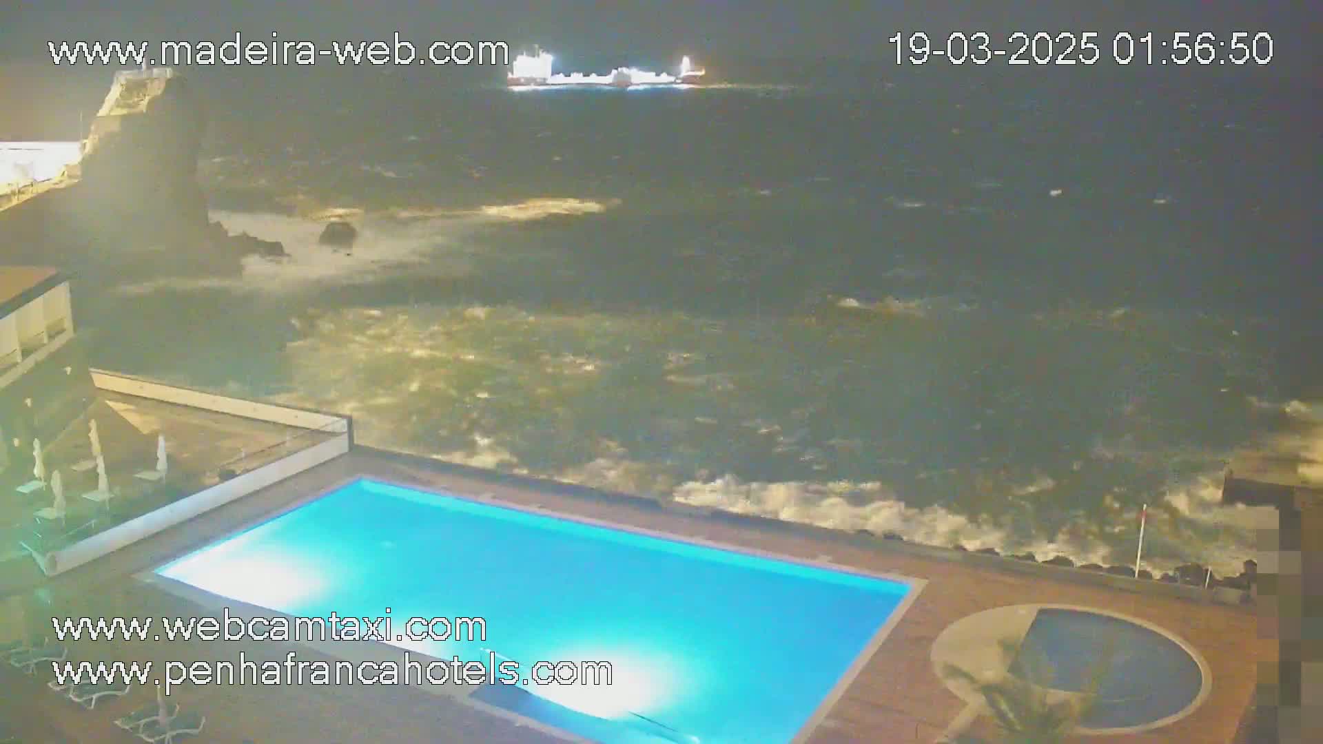 A nighttime view of a swimming pool at a resort overlooking a turbulent ocean with waves crashing against the shore.