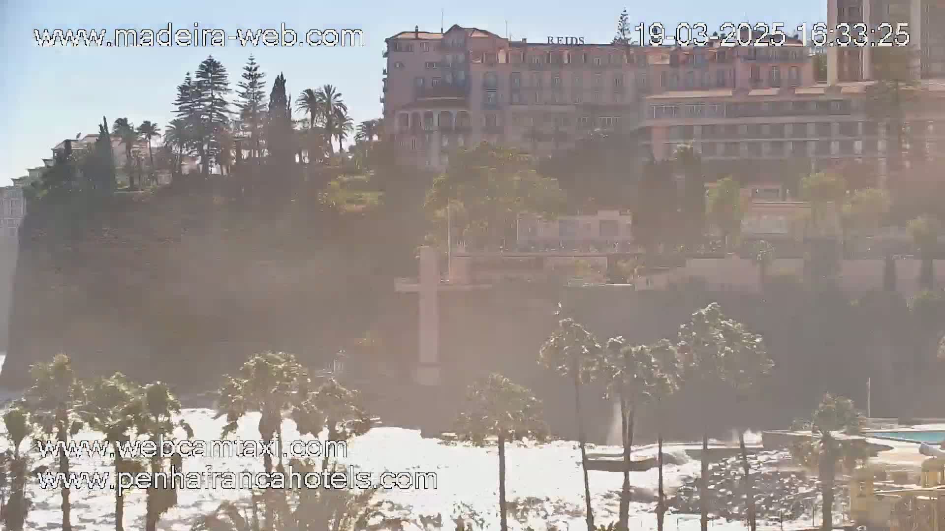 A hazy, sunny day reveals a coastal scene with a large building perched on a cliff overlooking a rocky shore with crashing waves.