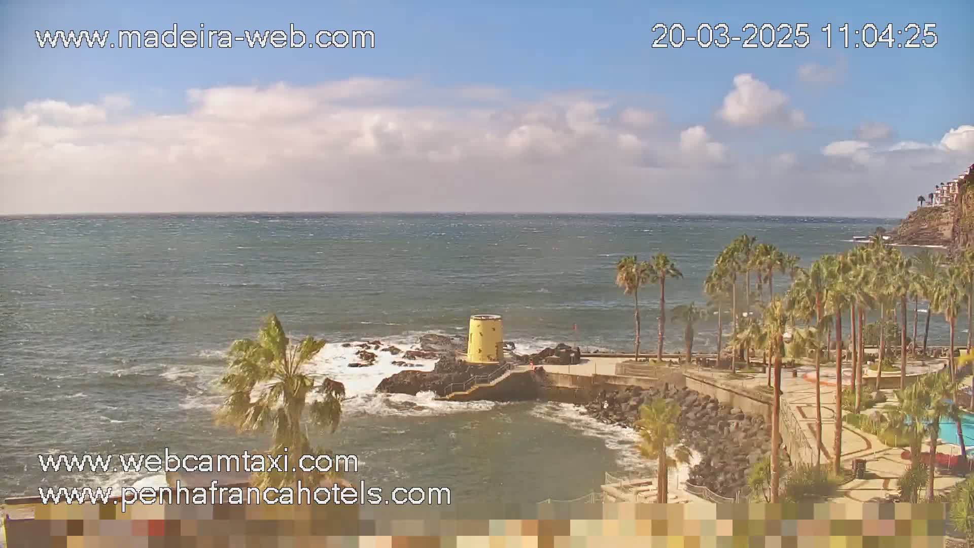 A partly cloudy day reveals a rocky coast with palm trees, a small yellow structure near the water, and a calm ocean with small waves.
