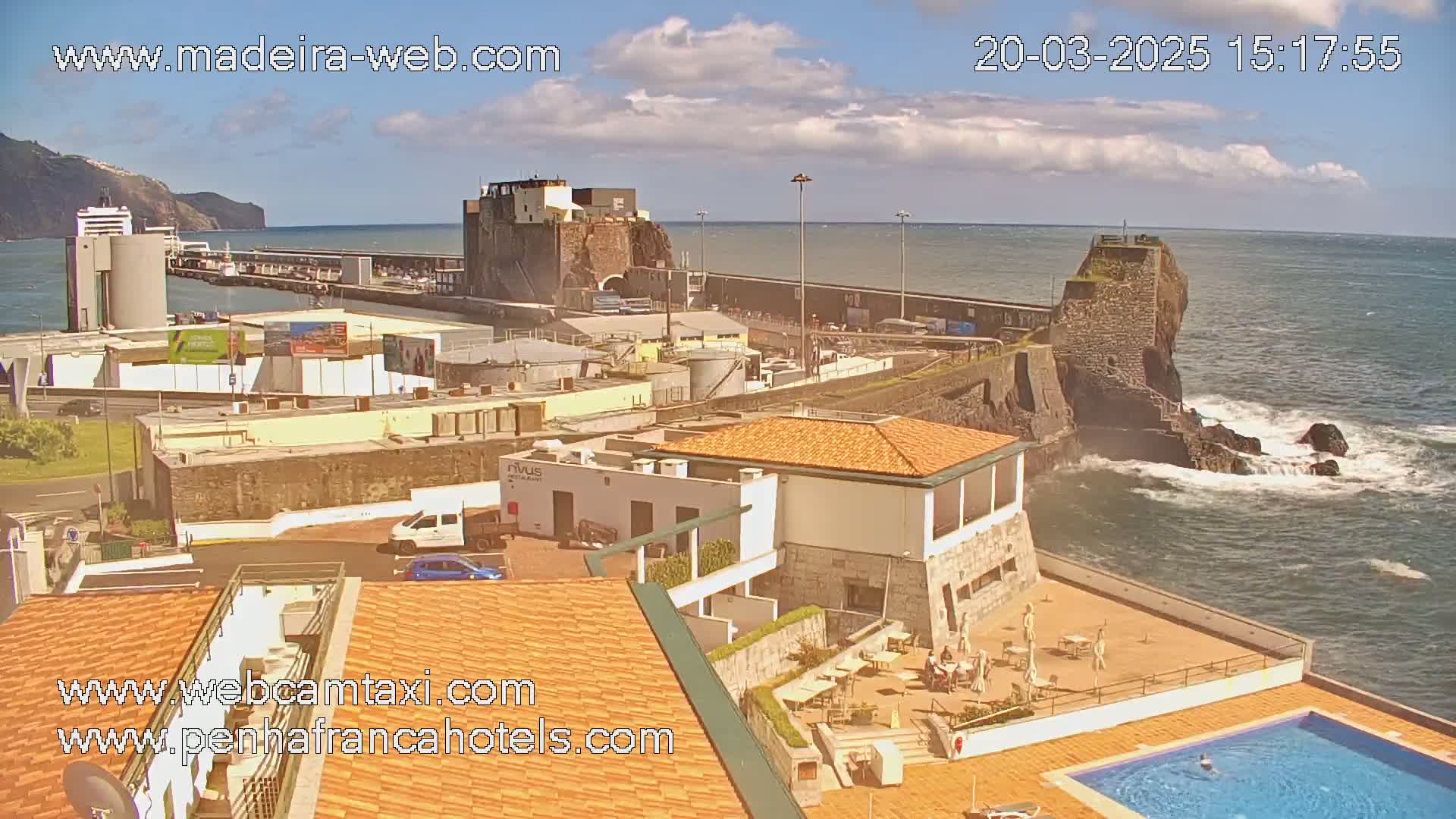A coastal scene on a partly cloudy day shows a harbor with buildings, a fortress on a cliff, and a hotel with a pool overlooking the ocean.