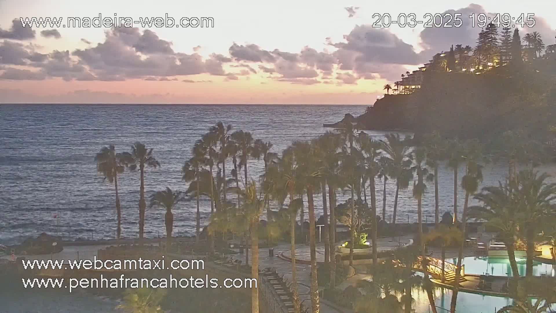 A coastal resort at sunset, featuring palm trees, swimming pools, and buildings situated on a cliff overlooking a calm ocean under a partly cloudy sky.