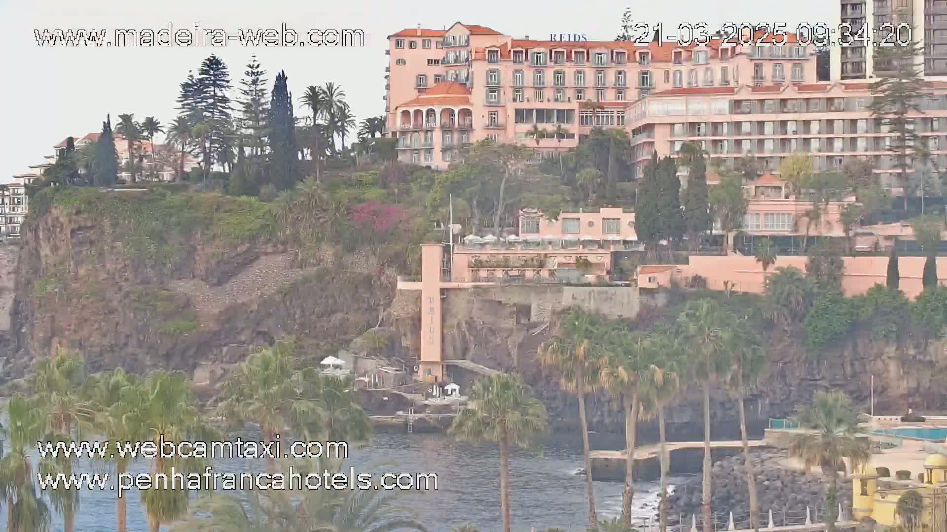 A pink hotel complex sits atop a dark cliff overlooking a calm ocean dotted with palm trees under an overcast sky.