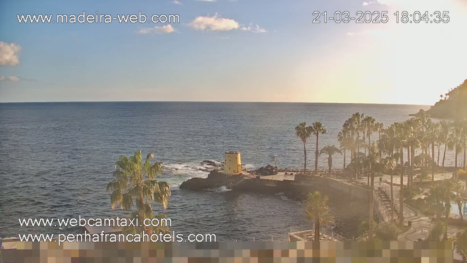 A calm ocean scene at sunset, featuring palm trees lining a rocky coast with a small yellow tower, under a partly cloudy sky.