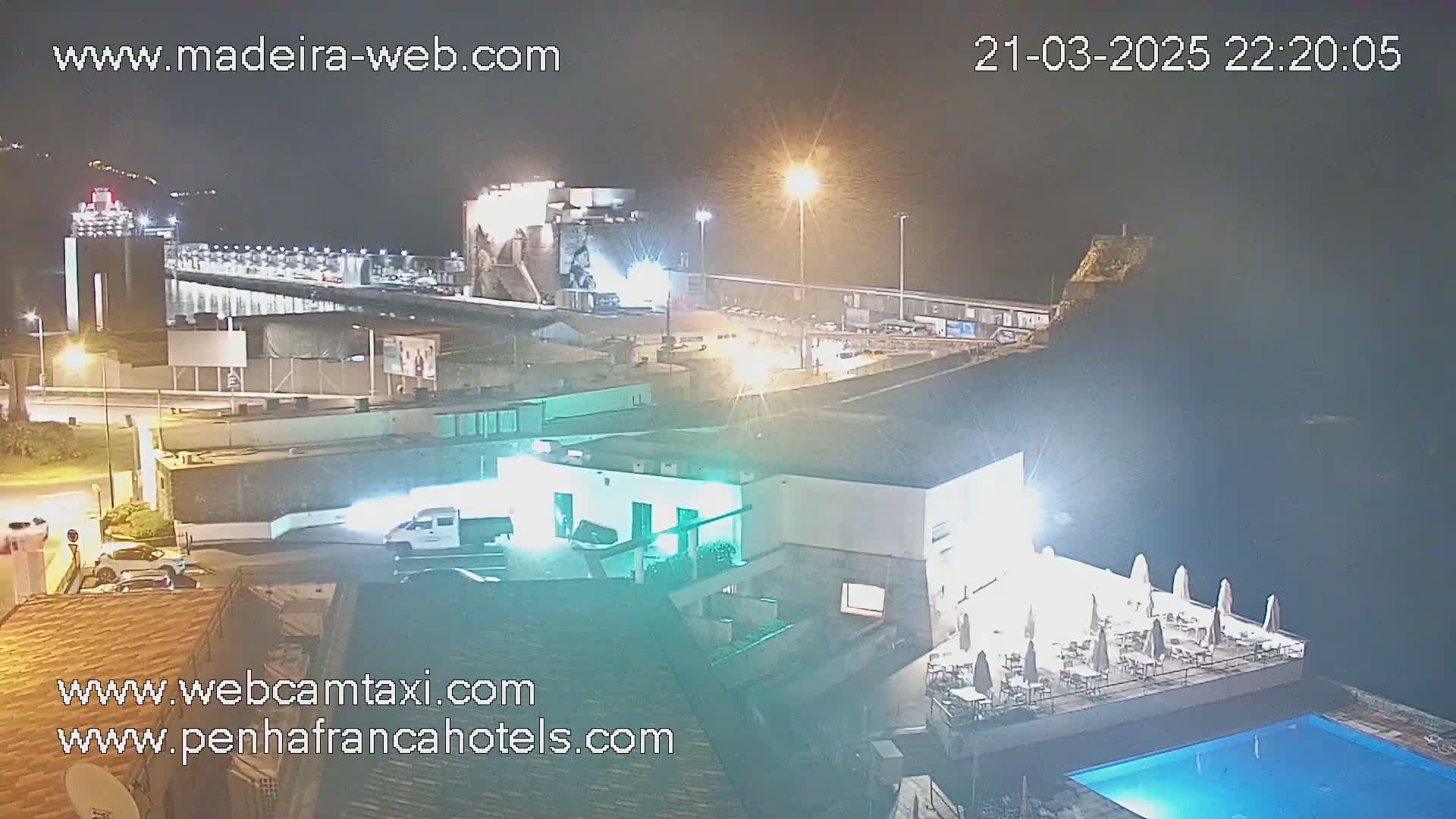 A nighttime view of a waterfront area with a building containing a pool and outdoor seating, lit by artificial lights, next to a large industrial structure under a clear, dark sky.