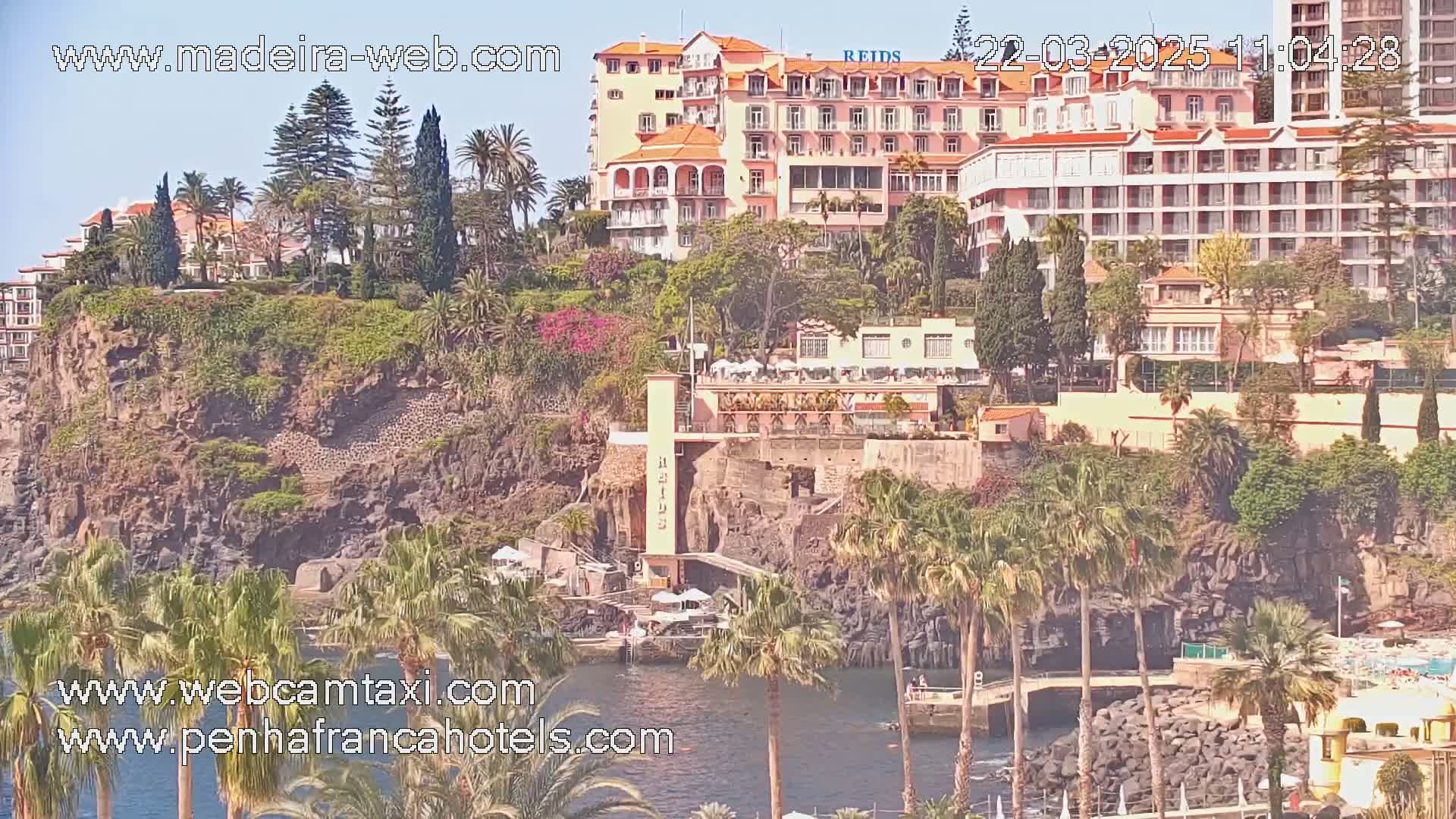 A coastal scene on a sunny day shows several hotels and buildings situated on a rocky cliff overlooking a calm body of water dotted with palm trees.