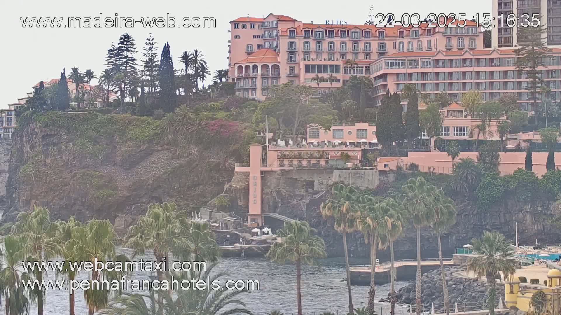 A large pink hotel sits atop a rocky cliff overlooking a calm body of water, surrounded by lush vegetation and palm trees under an overcast sky.