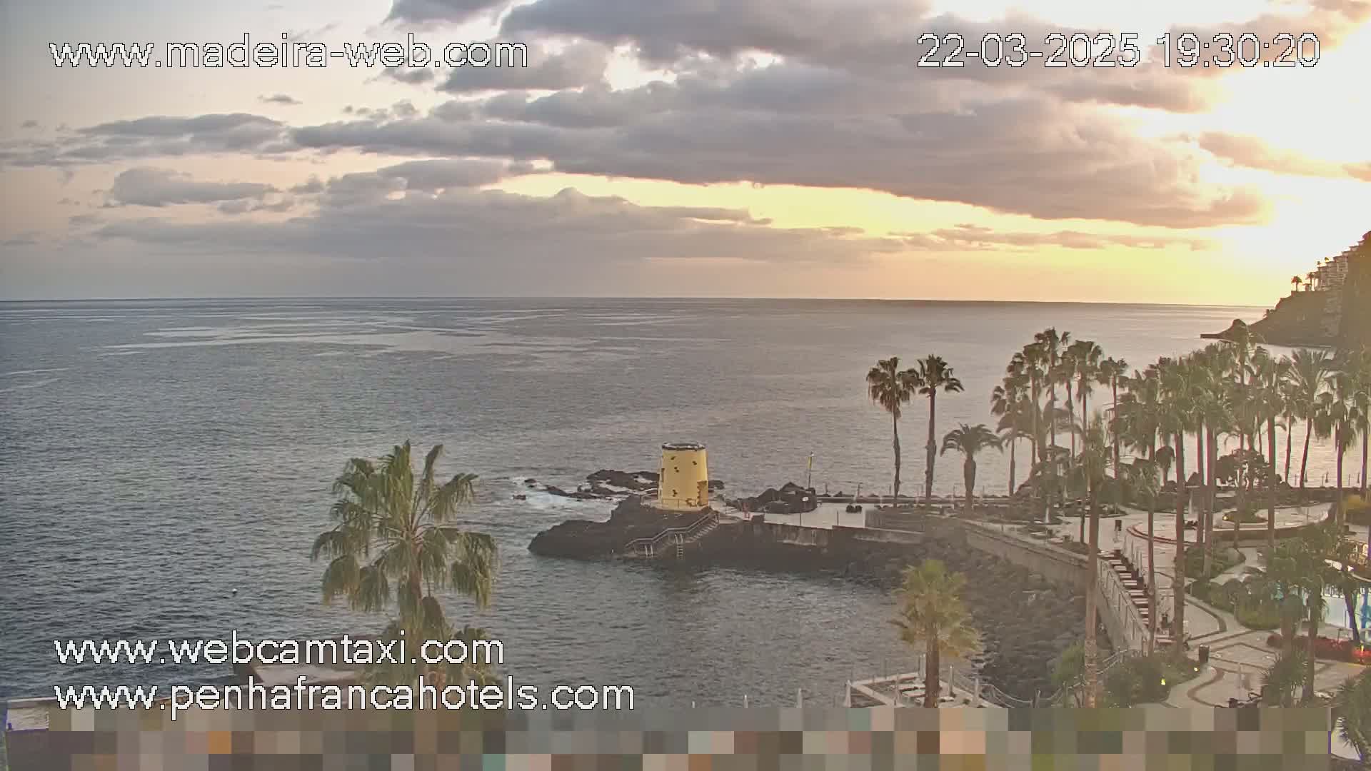 A partly cloudy sunset over a calm ocean, with palm trees and a small yellow tower on a rocky outcrop near a resort.