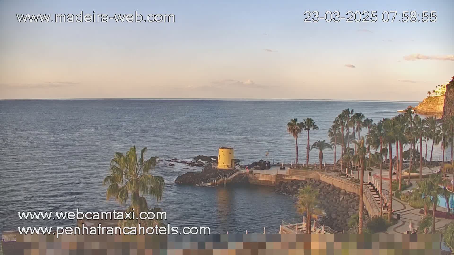 A calm ocean under a mostly clear sky is seen from an elevated vantage point, showing palm trees and a small yellow structure on a rocky shoreline.