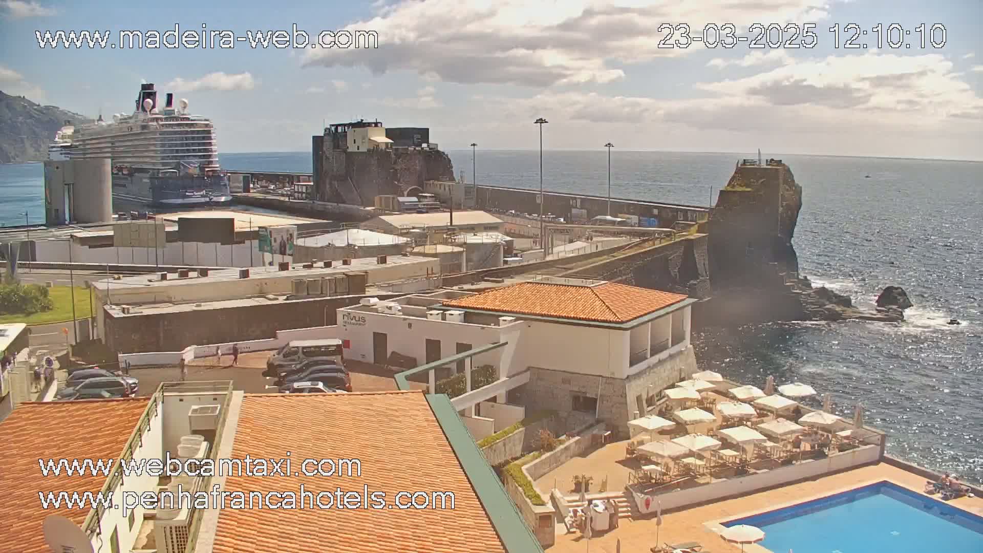 A cruise ship is docked near a coastal town with a swimming pool and patio umbrellas under partly cloudy skies.