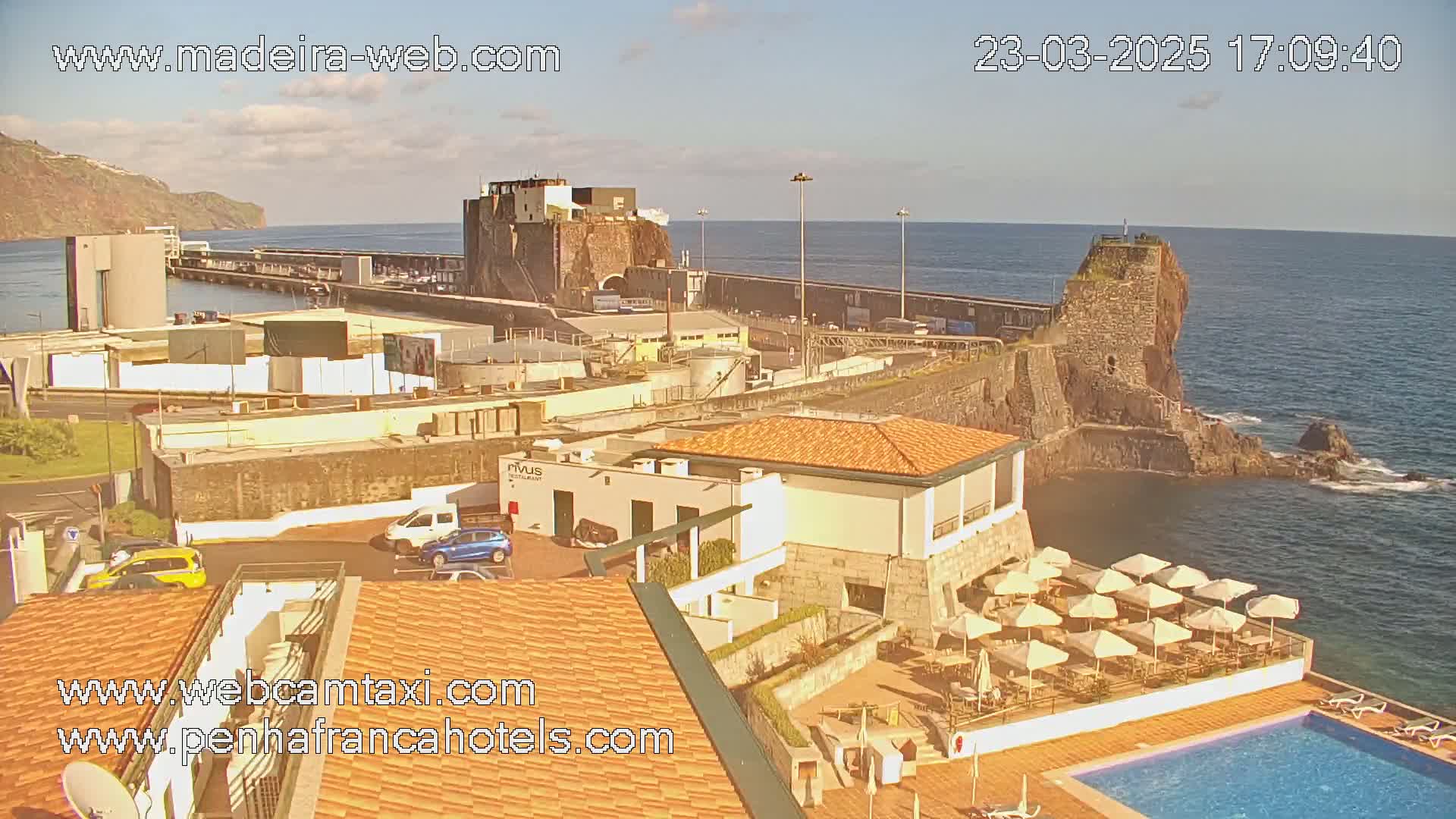 A coastal scene under sunny skies shows a harbor with stone structures, a building with outdoor seating and a swimming pool, and a rocky shoreline extending into a calm ocean.