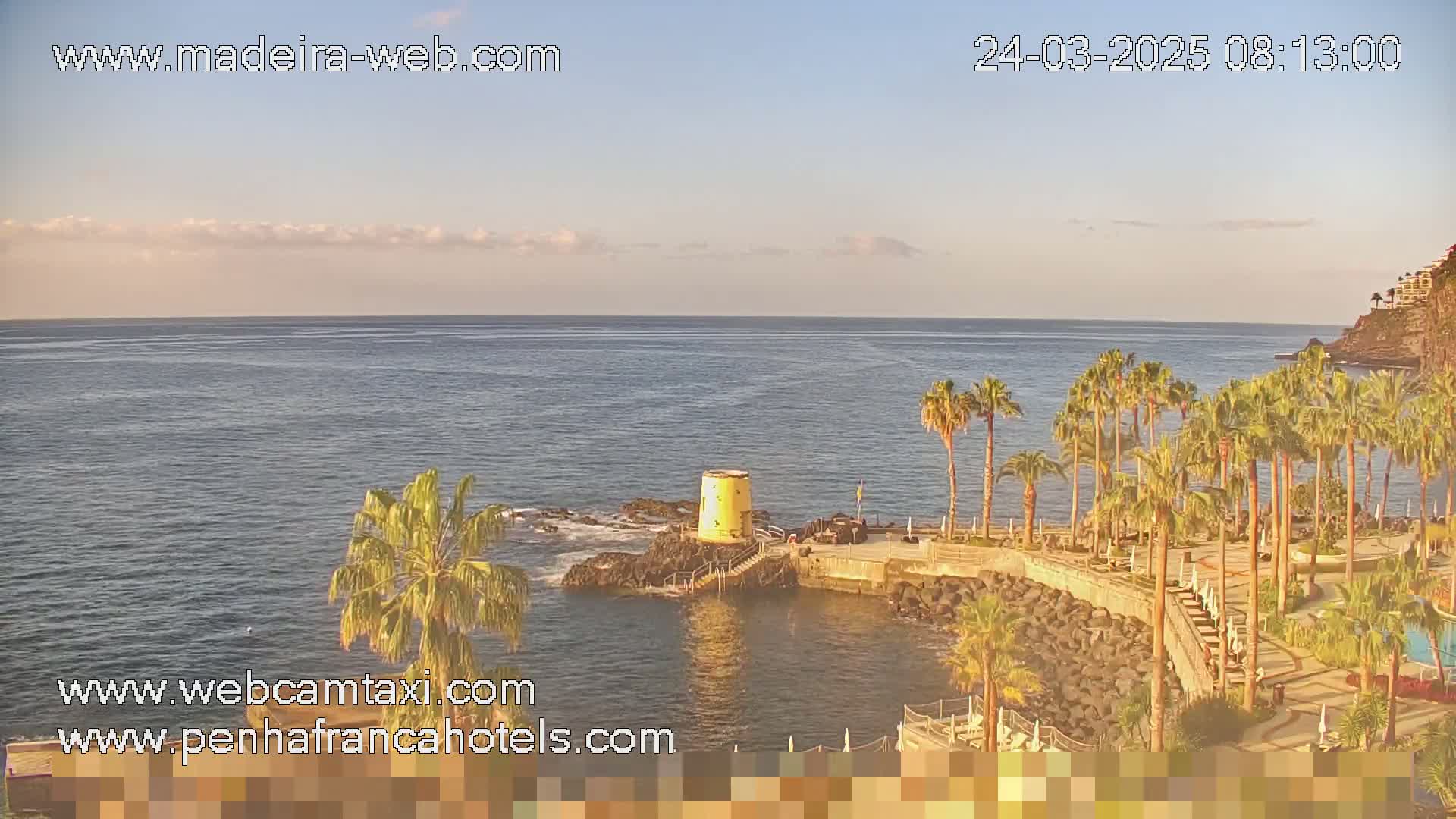 A calm ocean is seen from a coastal vantage point featuring palm trees and a small yellow structure on a rocky outcrop under a mostly clear sky.
