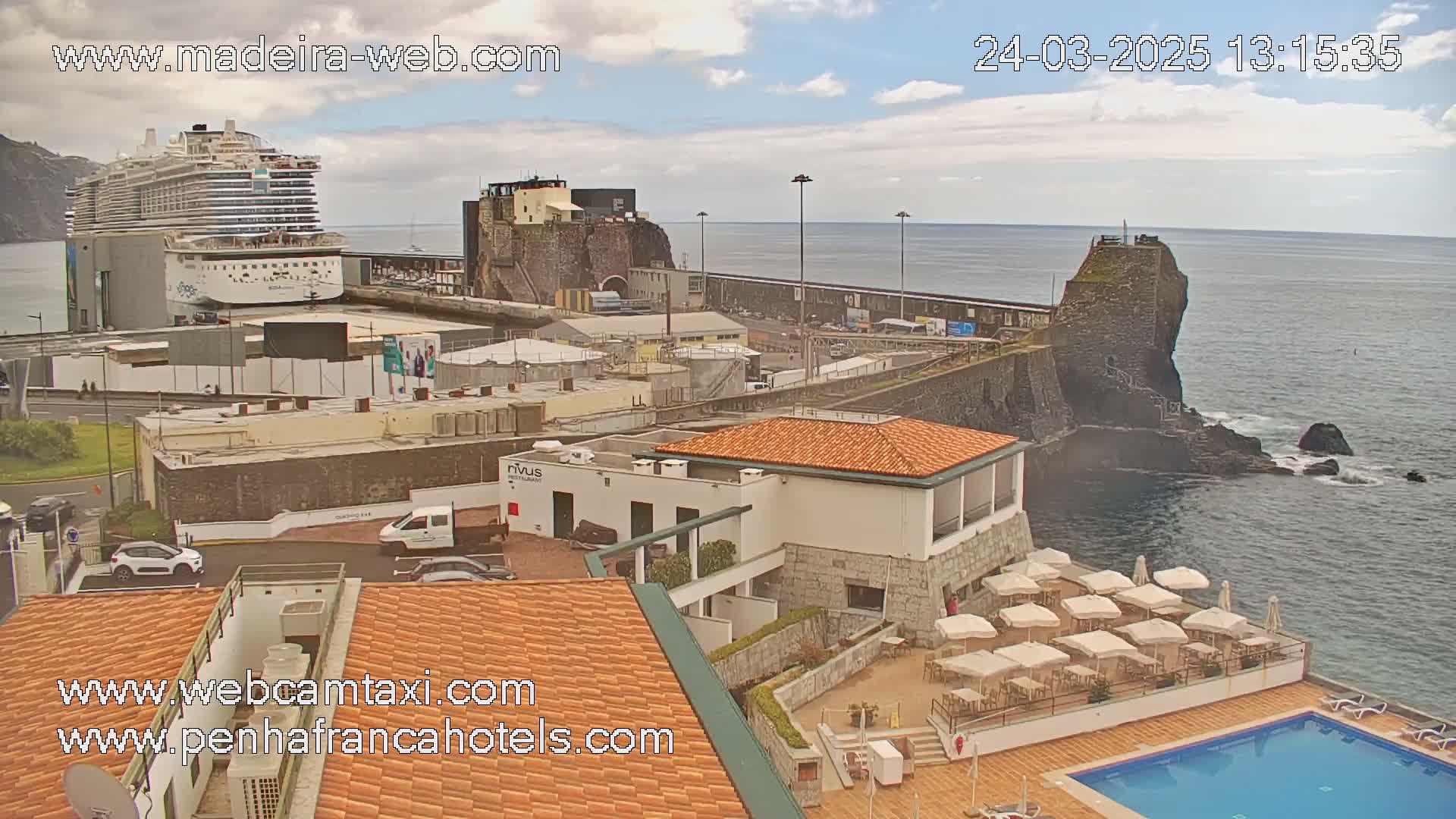 A cruise ship is docked at a port next to a rocky coastline with a building and outdoor pool, under partly cloudy skies.