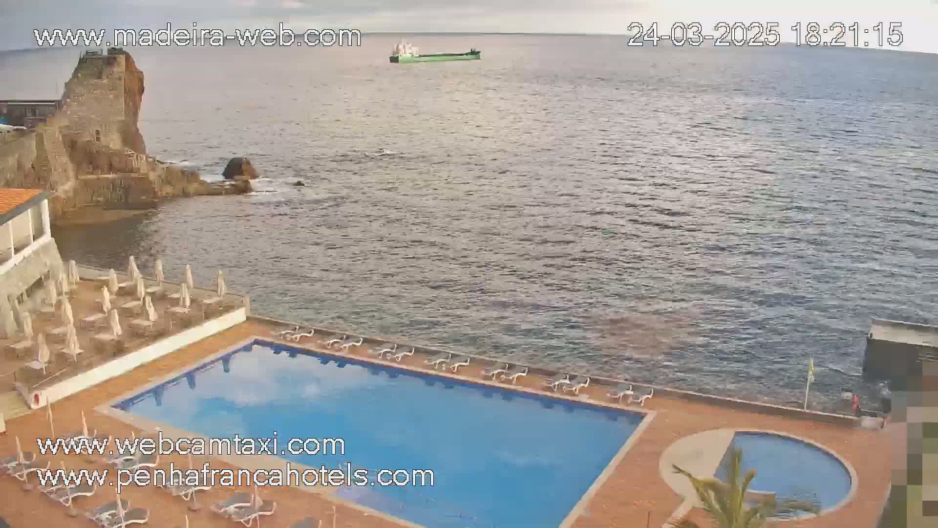 A hotel pool area with lounge chairs and umbrellas overlooks a calm ocean with a distant ship, under an overcast sky.