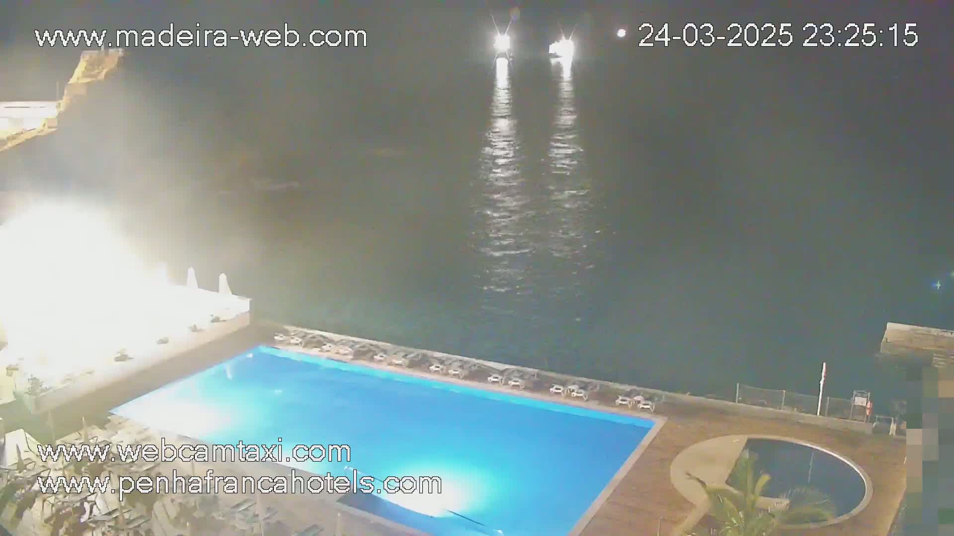 A nighttime view shows a swimming pool and a smaller round pool at a resort overlooking calm ocean water with two distant boats visible.