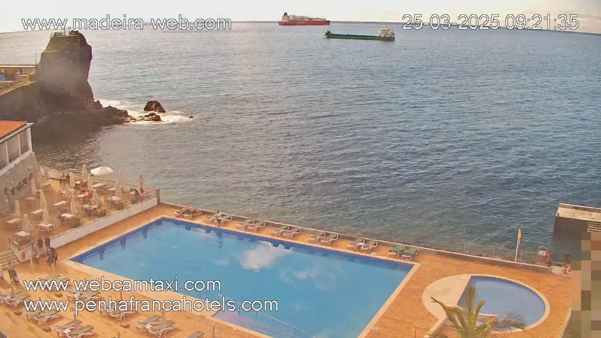 An outdoor swimming pool and patio area at a seaside hotel are shown, with two ships visible on a calm ocean in the background.