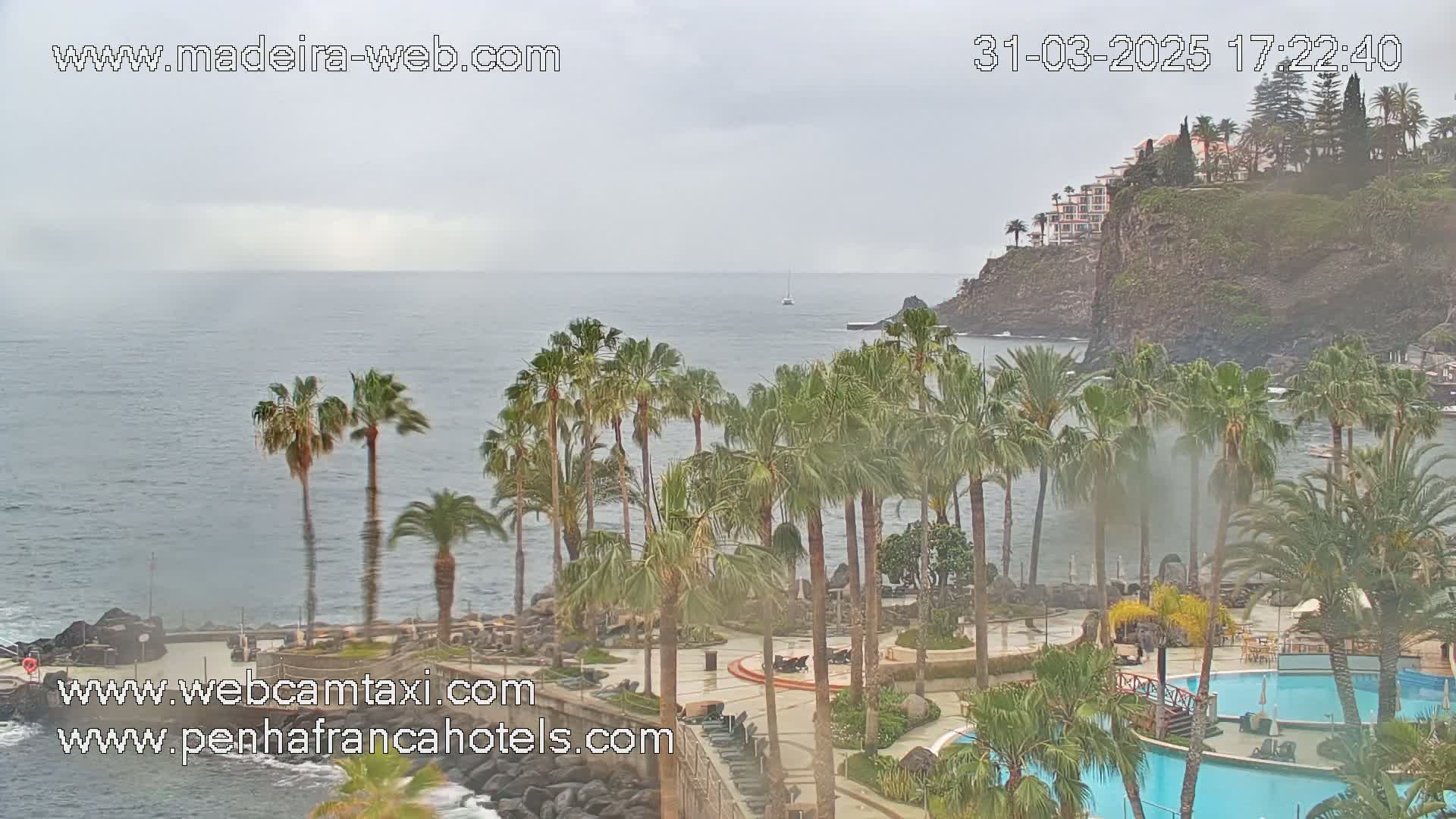 A hazy, overcast day shows a resort with palm trees and swimming pools overlooking a rocky coastline and ocean, with a sailboat in the distance.
