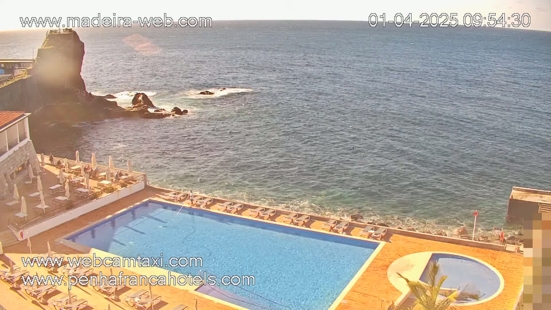A resort pool and patio area overlooks a rocky coastline and ocean under sunny skies.