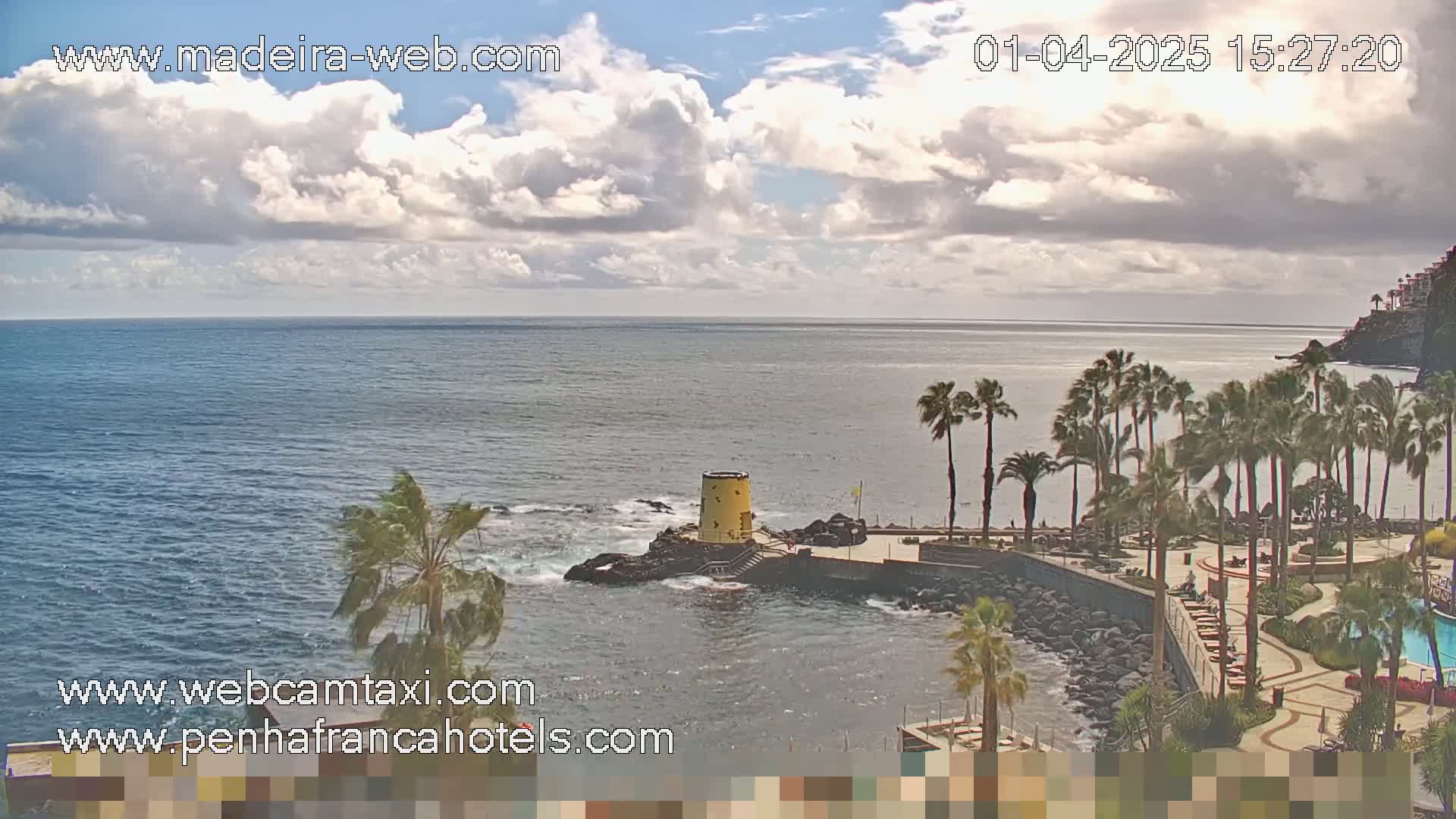 A partly cloudy day reveals a rocky coastline with palm trees, a small yellow tower, and a swimming pool overlooking a calm ocean.