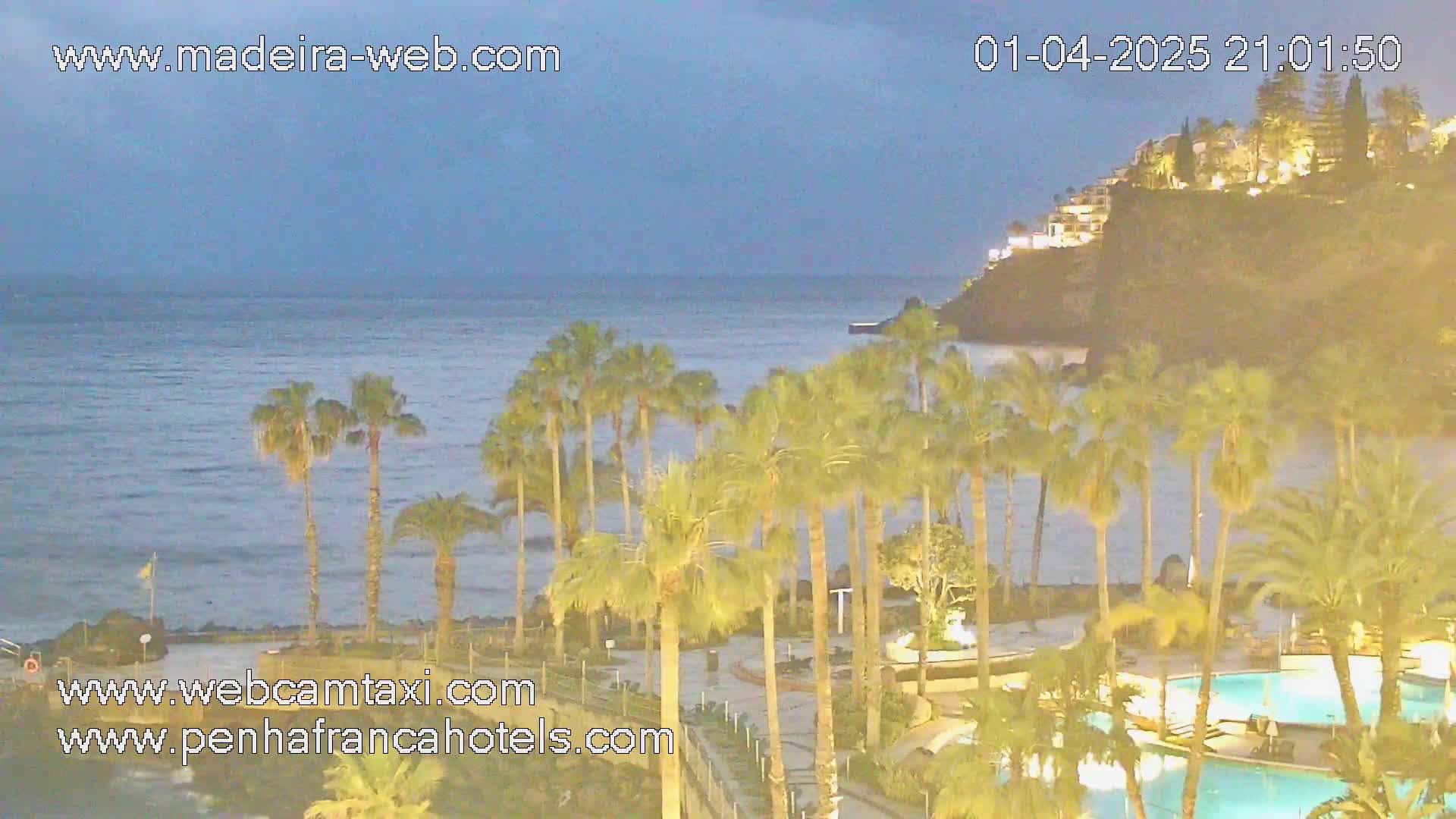 A nighttime view of a calm ocean, palm trees lining a resort area with illuminated swimming pools, and a cliffside with buildings in the background under a dusky sky.