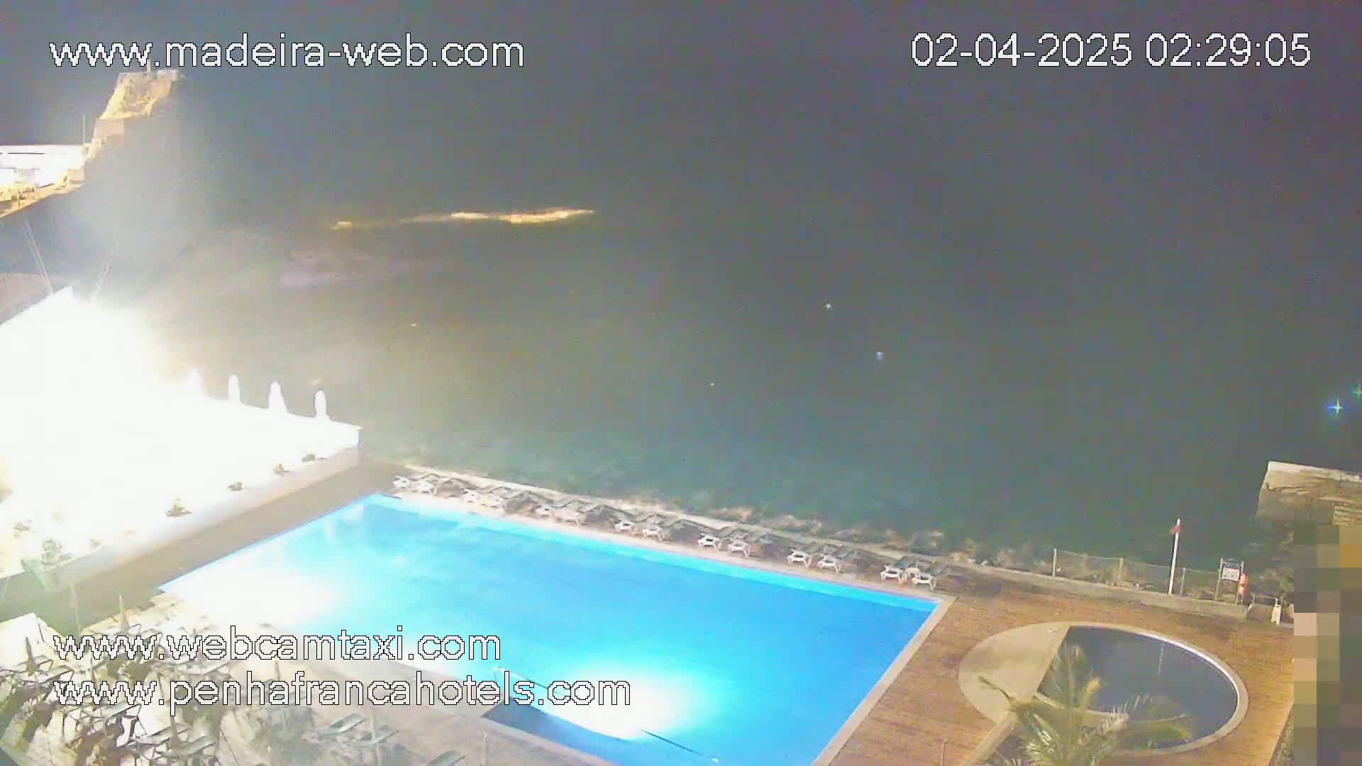 An outdoor nighttime scene shows a large, illuminated swimming pool next to a smaller, round pool, overlooking a dark ocean.